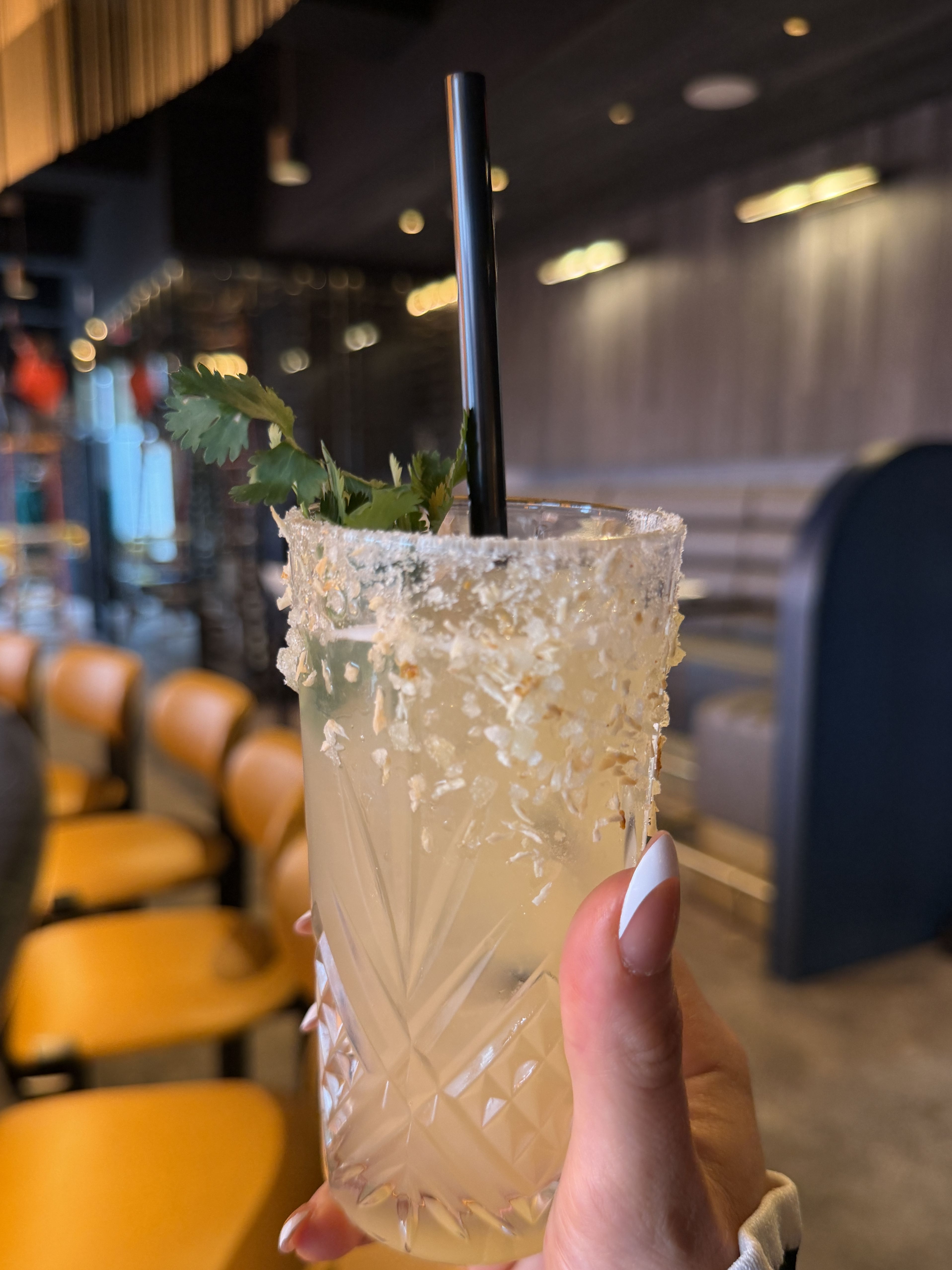 Close-up of a margarita-style cocktail with a salt-crusted rim in a crystal glass, a black straw and a mint sprig, held by a hand. Warm, blurred bar interior with yellow chairs.