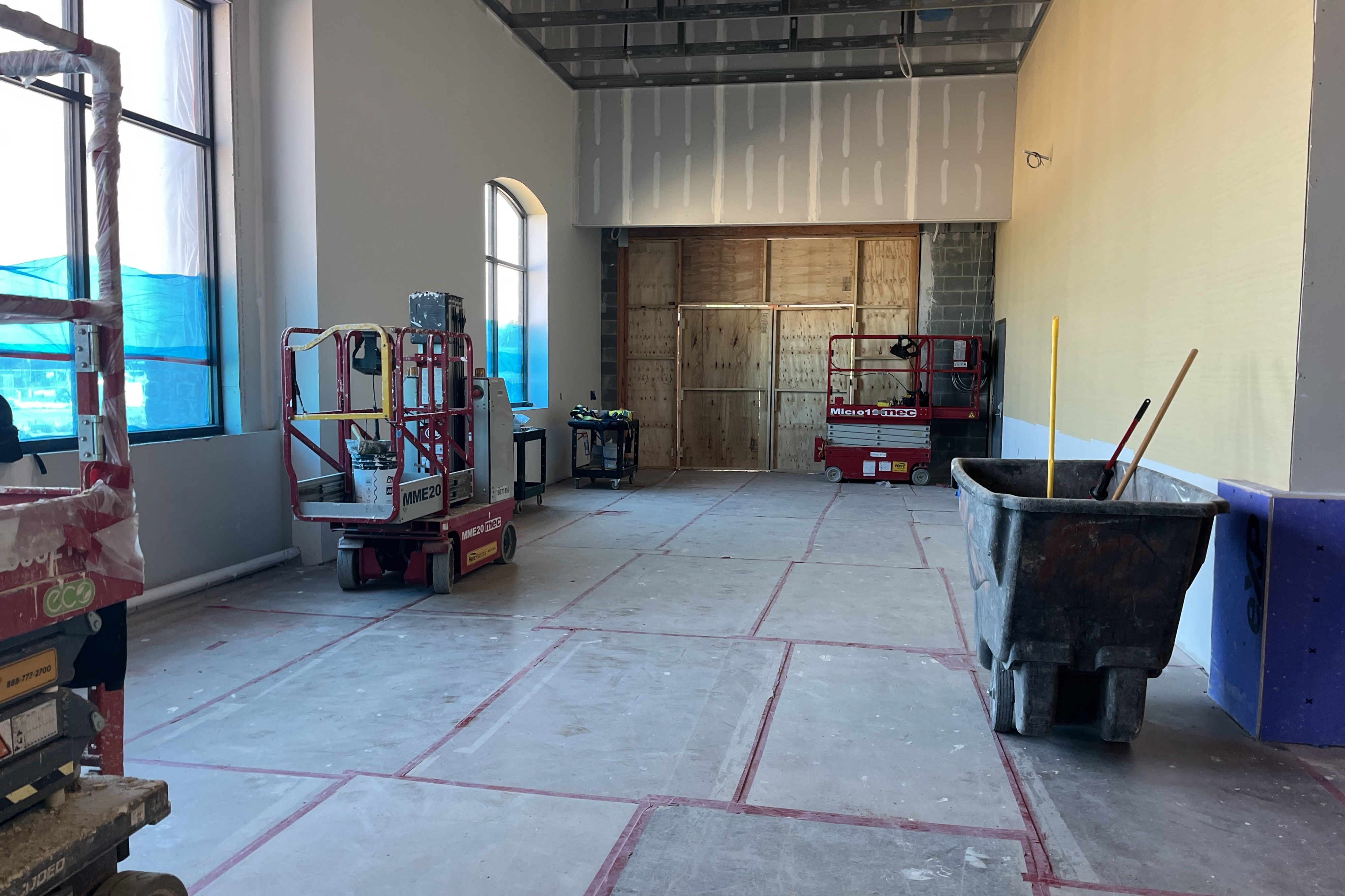 Interior construction site with blue window film, red scissor lifts and scaffolding, plywood back wall, drywall panels, and a yellow wall on the right; a large dumpster in the foreground.