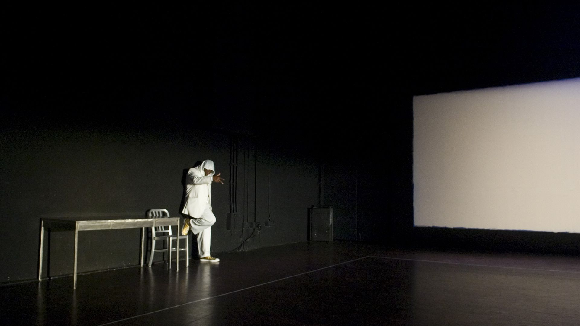 A man wearing all white leans against a table near a big blank screen in a black room