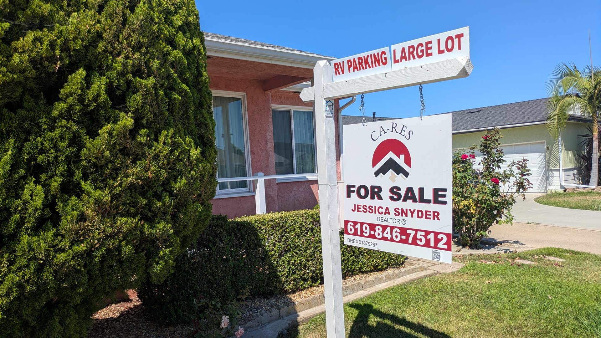 Sunny suburban scene with a pink stucco house behind dense green hedges. A white post holds a sign reading "RV PARKING LARGE LOT" beside a "FOR SALE" sign for Realtor Jessica Snyder.