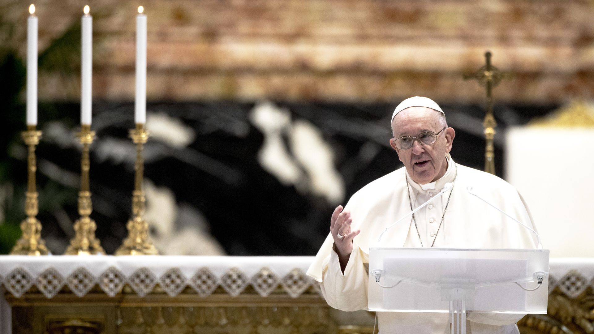 Pope Francis speaks while standing behind a podium, several tall candles stand behind him