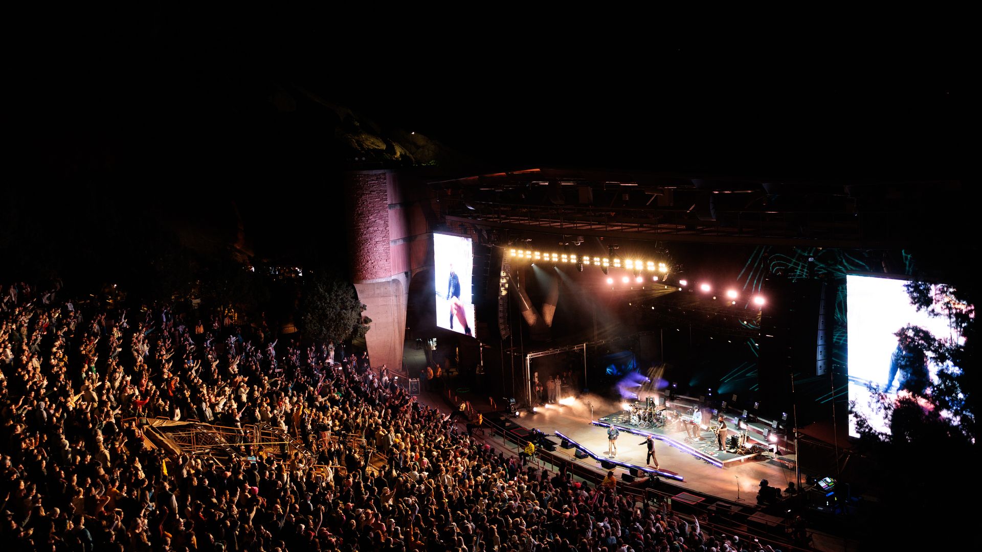 Nighttime outdoor concert in a large stadium with a packed audience. Bright stage lights and large screens flank the stage, performers visible at center amid fog and colorful lighting.
