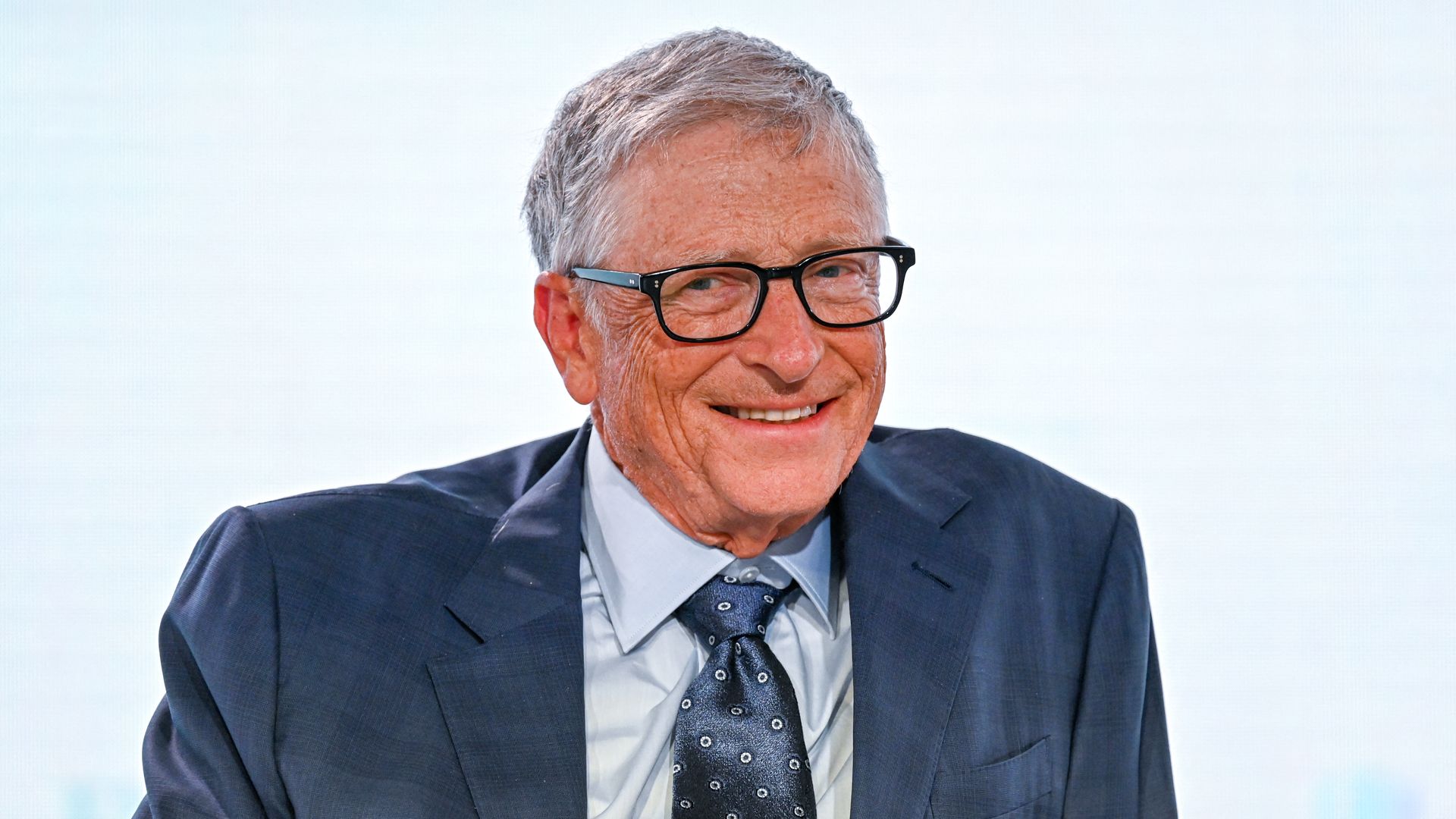 A person in black rimmed glassed and a dark blue suit smiles in front of a light gray background
