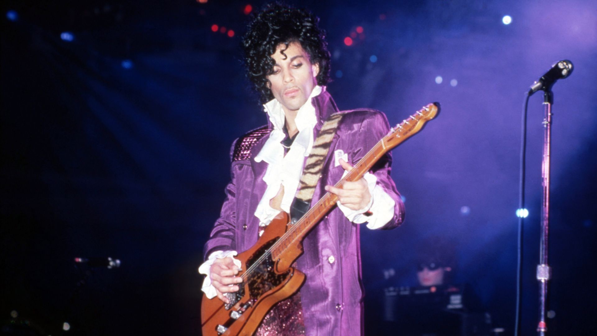 A musician on stage wearing a purple velvet jacket with a white ruffled shirt, playing a bass guitar. Blue and purple stage lighting and a microphone stand to the right.
