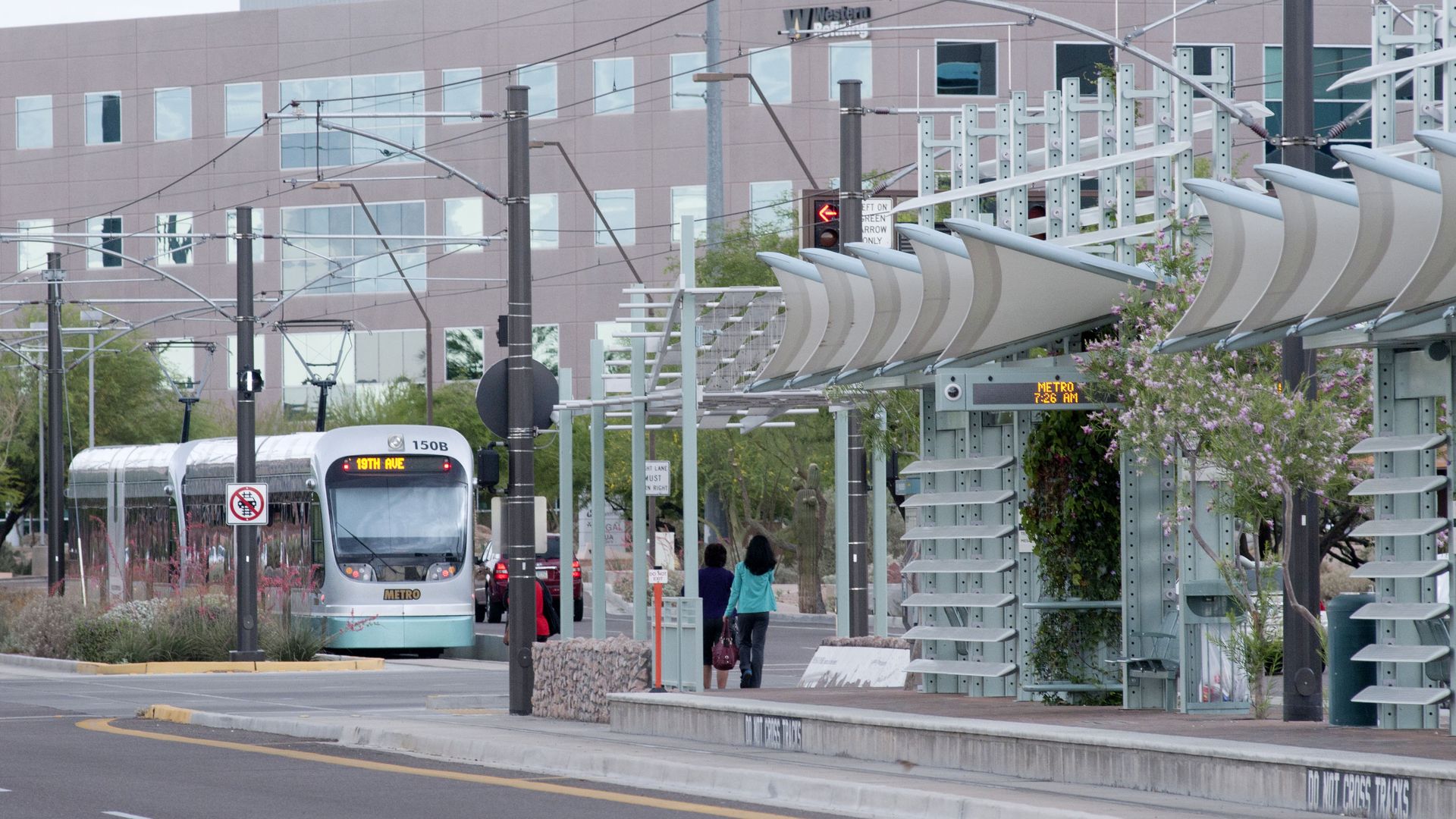 A silver light rail train pulls up to a platform with a row of fin-like awnings over it, with two people waiting on the platform. 