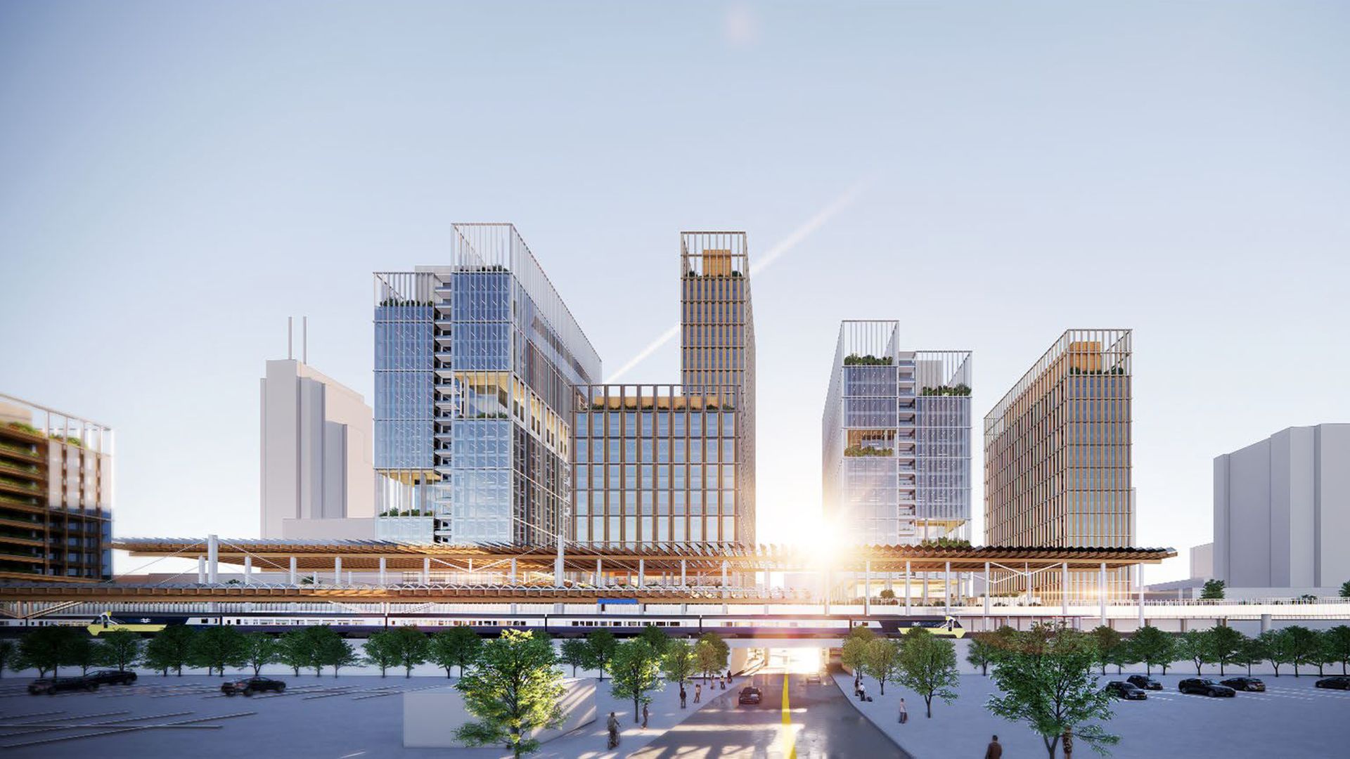 Modern cityscape at sunrise with tall glass-covered buildings surrounding an elevated train station, trees lining the streets, and cars and people below.