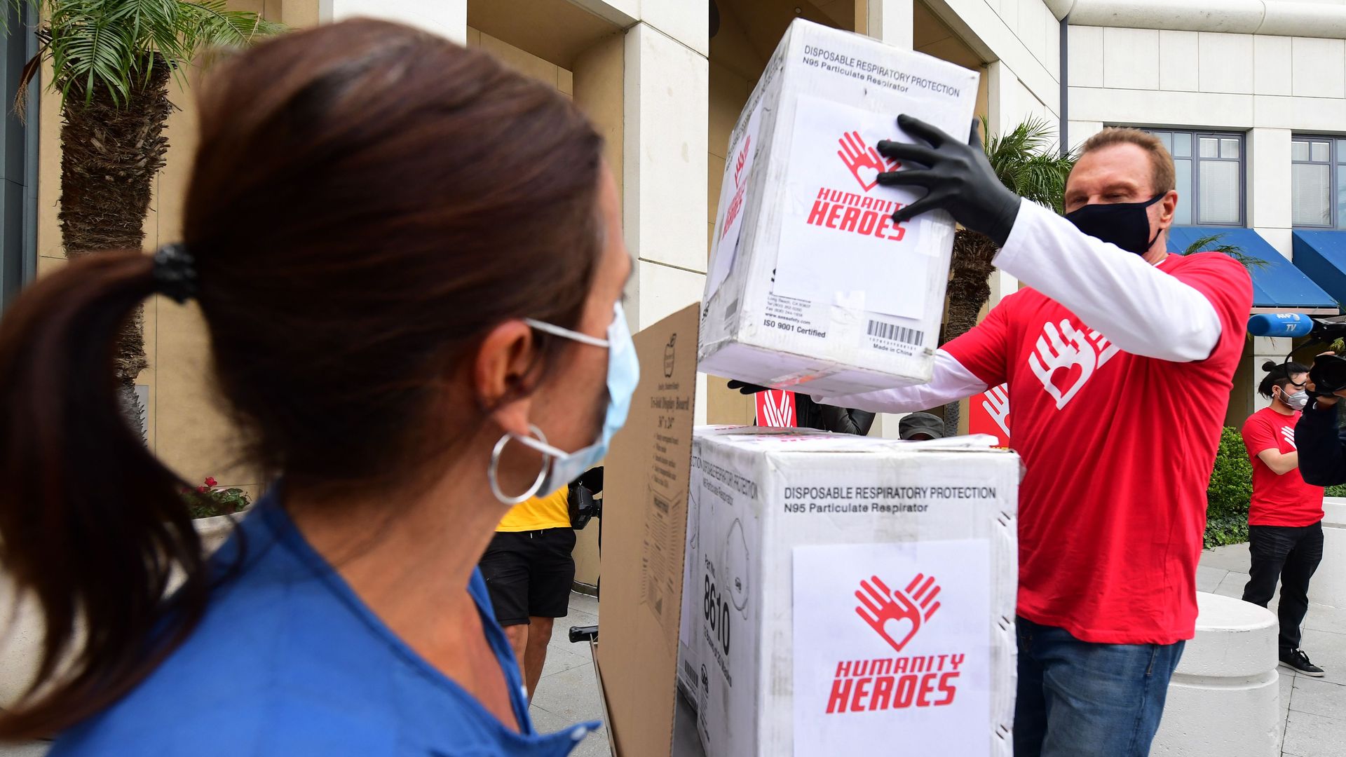 In this image, a man stacks boxes while a woman wearing a face mask watches