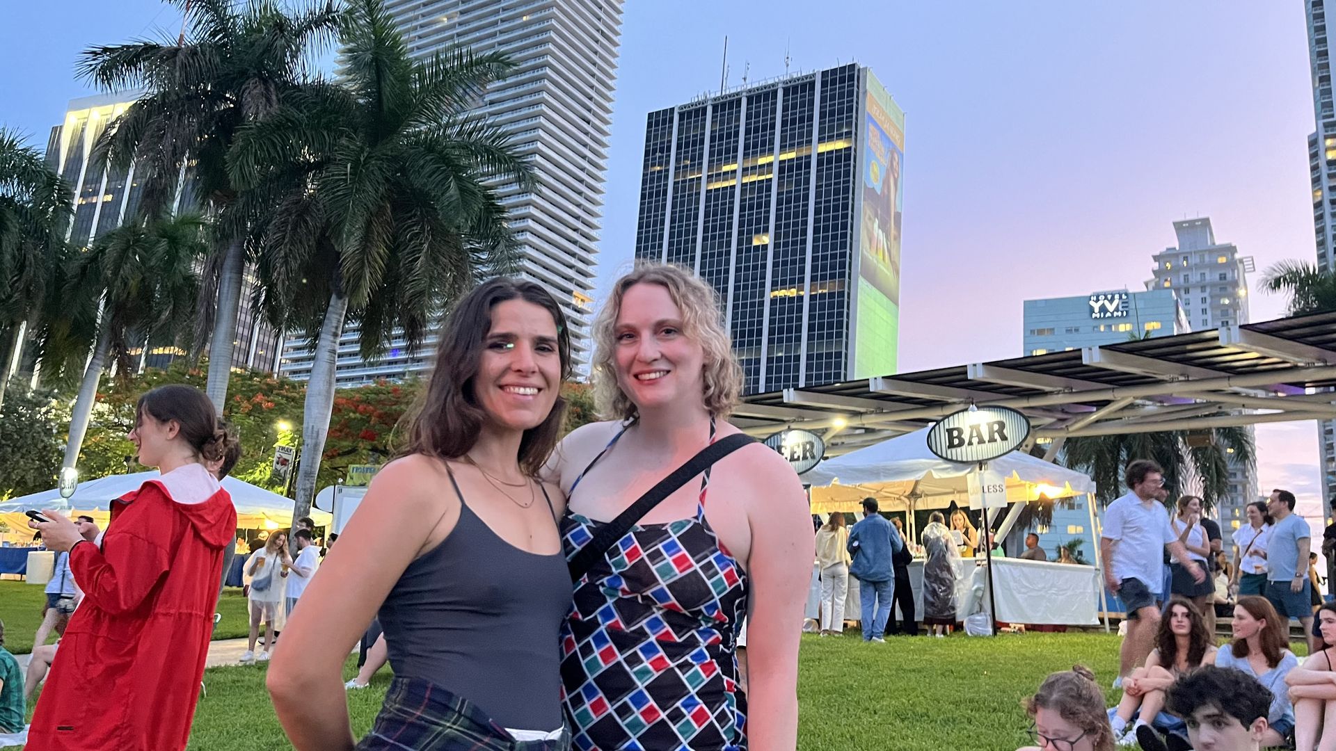 Two women, one in a gray tank and one in a plaid halter dress, smiling for a photo in front of several tall buildings and a blue-and-pink sky.