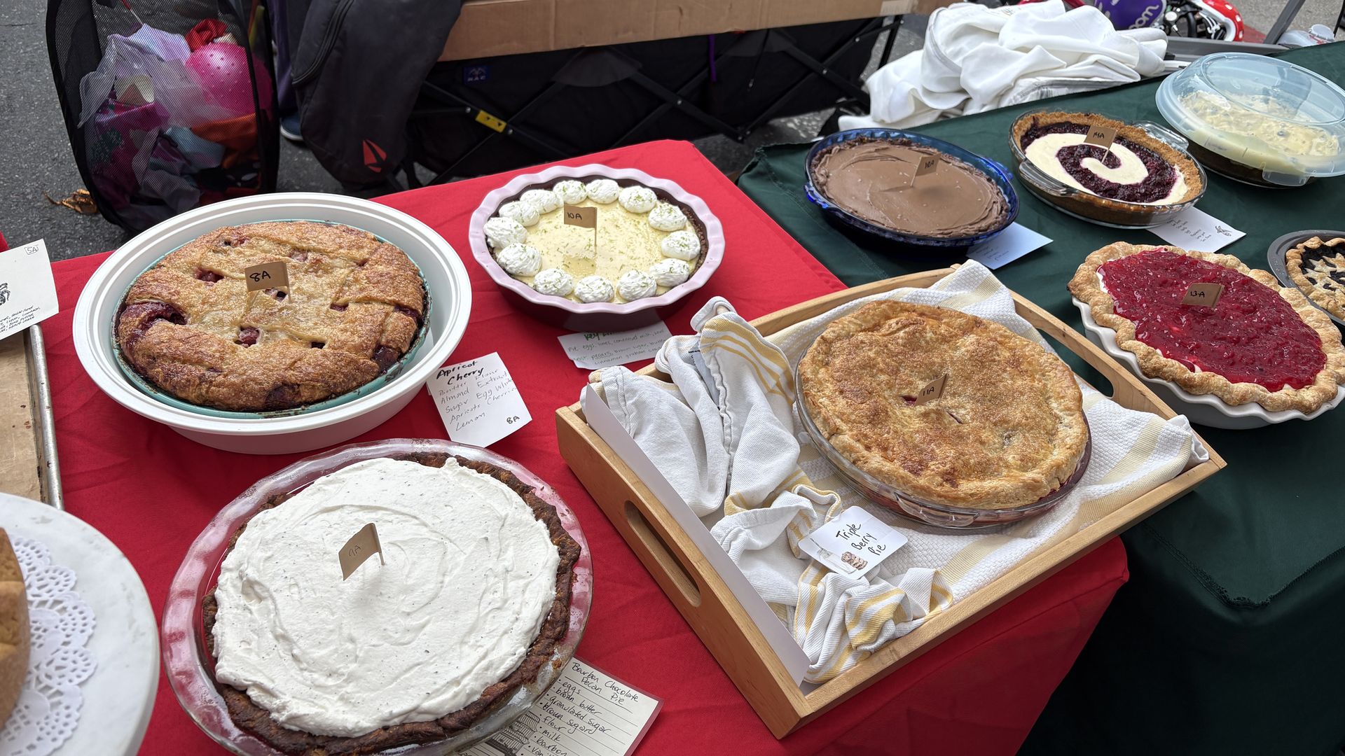 Assorted pies on a red-and-green table at an outdoor bake sale: lattice berry pie, lemon tart with whipped cream, chocolate pie, and a swirl berry pie; background shows boxes and a chair.