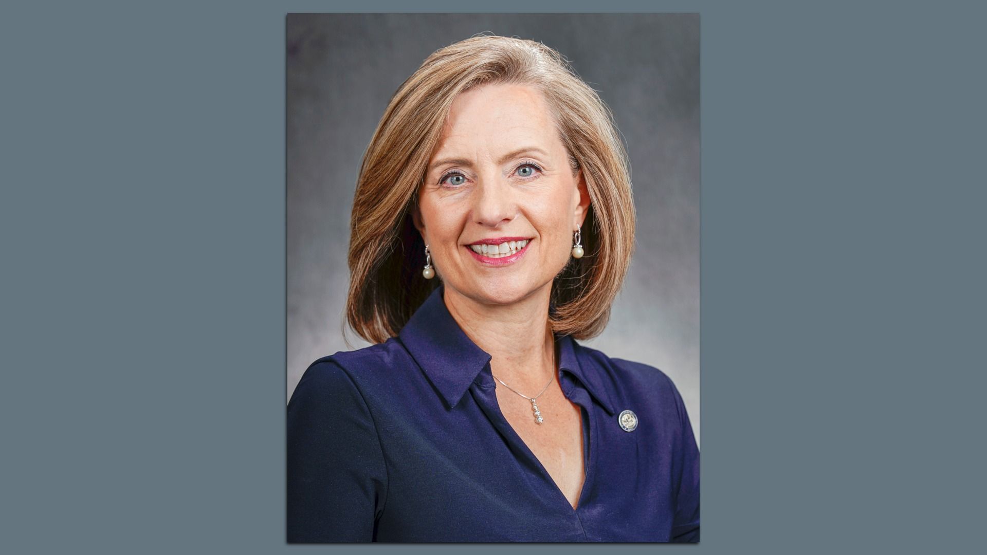 Portrait of a smiling woman with shoulder-length blonde hair, wearing pearl earrings, a silver necklace, and a dark blue blouse with a pin on the collar, against a gray background.