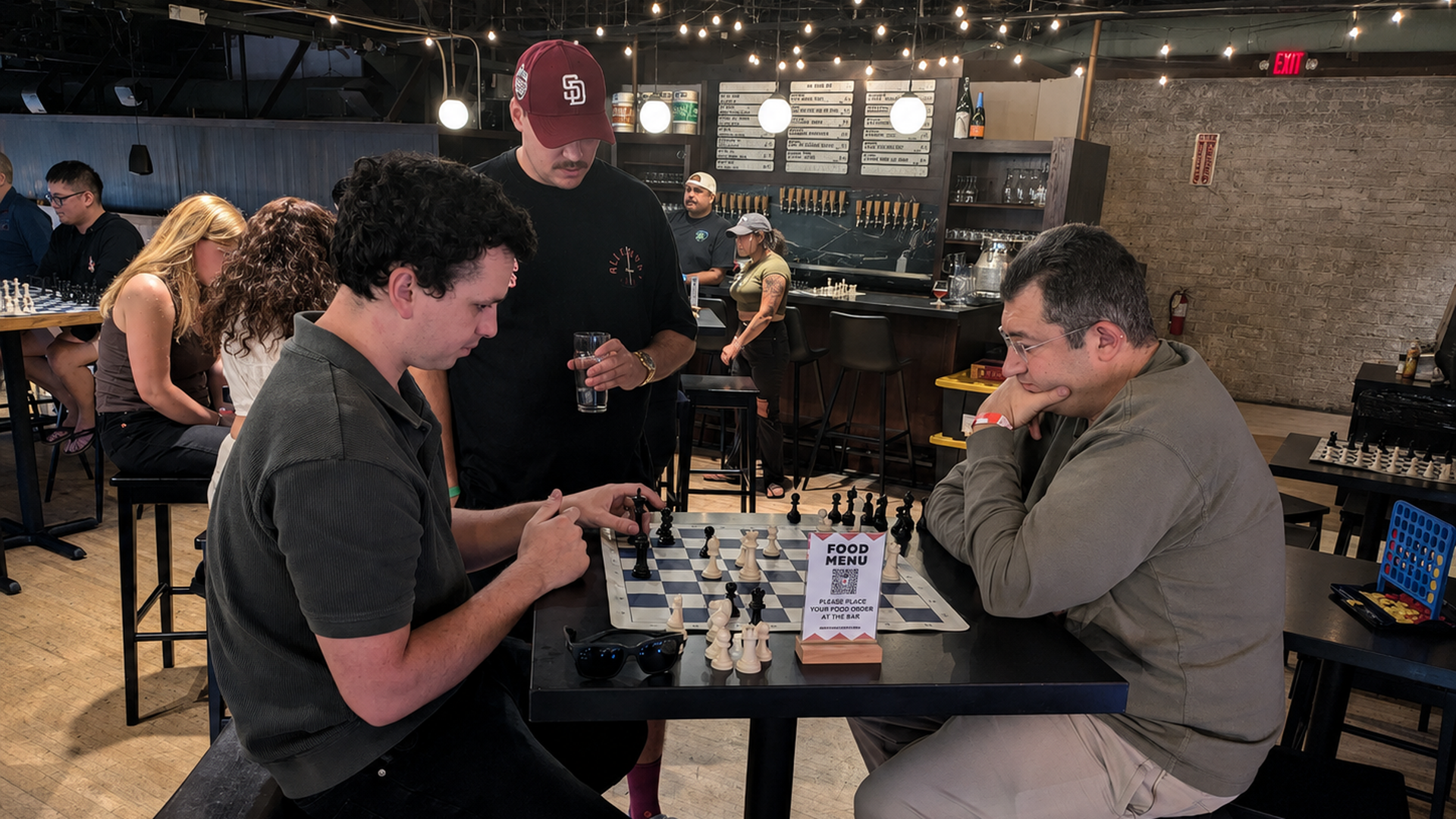 Two men play chess at a black cafe table. The left player studies the board while the right leans in. A man in a red cap watches from behind, with string lights above and a menu board nearby.