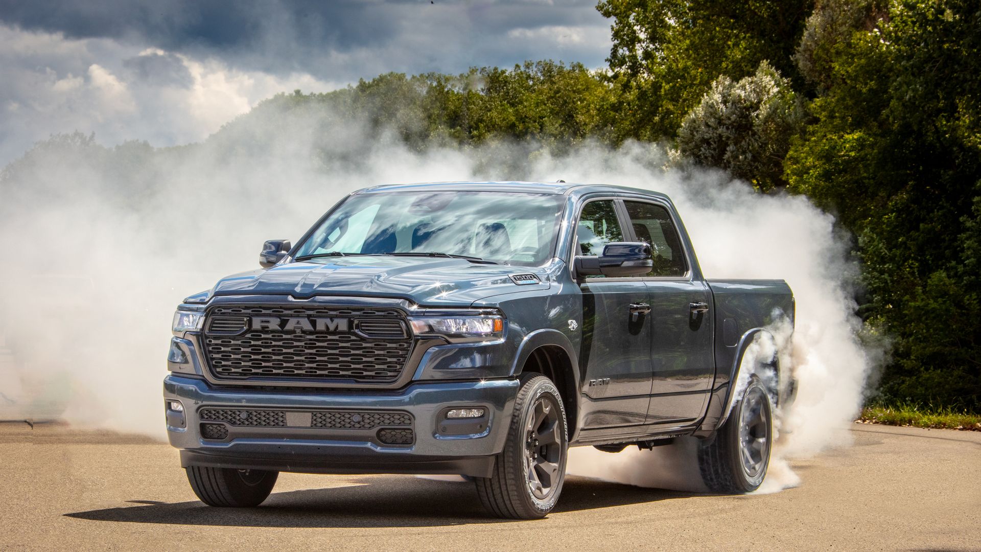 Gray RAM 1500 pickup truck with Hemi V8 engine performing a burnout, creating thick white smoke around the rear tires on a sunlit road with green trees in the background.