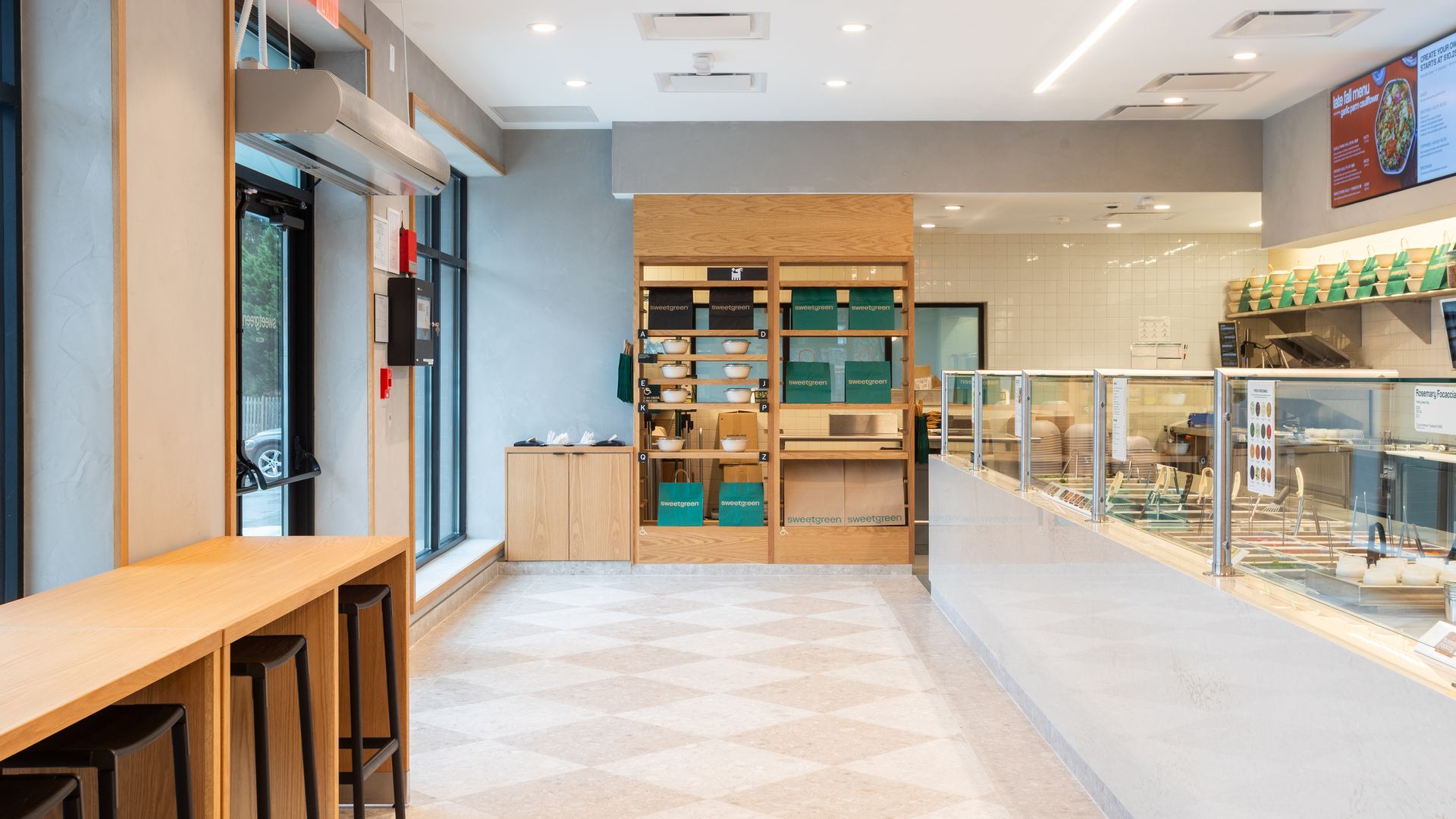 Bright cafe interior with a long wooden counter and stools on the left, tall windows; center shelves hold bowls and teal sweetgreen boxes; glass display case on the right and tiled wall with menus.
