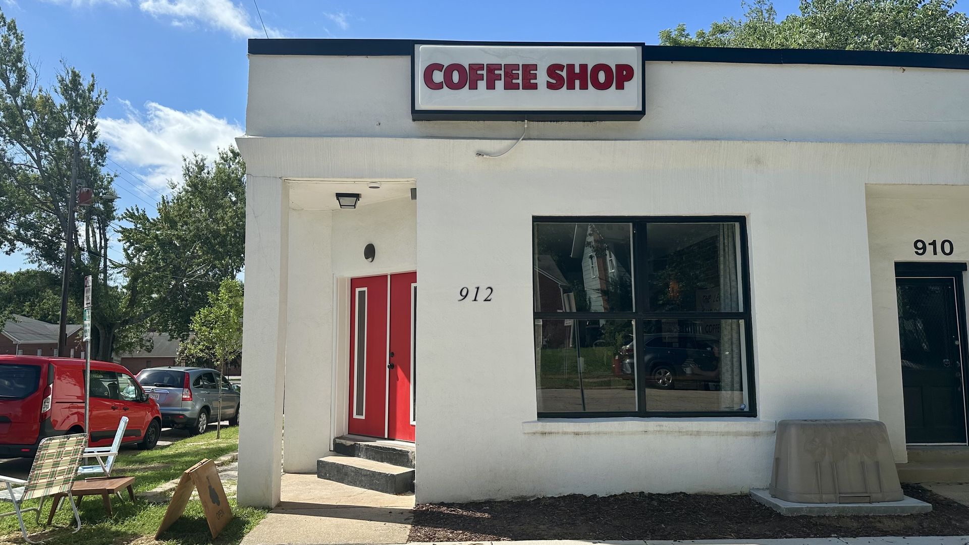 White, single-story coffee shop with a bold red COFFEE SHOP sign above a red door. Large front window; numbers 912 and 910 on the wall; outdoor chairs and parked cars under a blue sky.