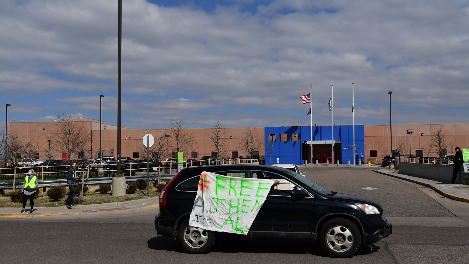 Black car with a large sign reading "#FREE THEM ALL Abolish ICE" in bright green and red letters, parked in front of a building an American flag and other flags. 