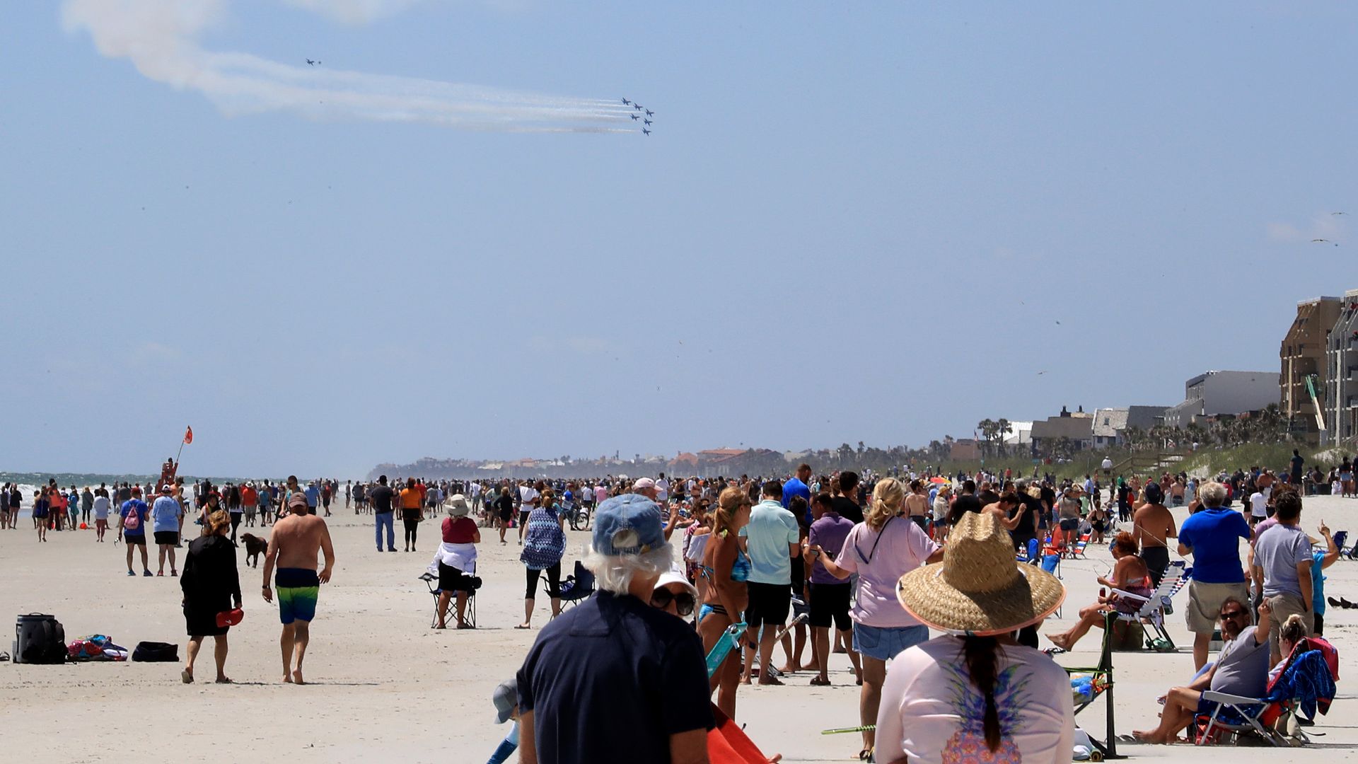 A crowd on a beach watches the Blue Angels fly overhead