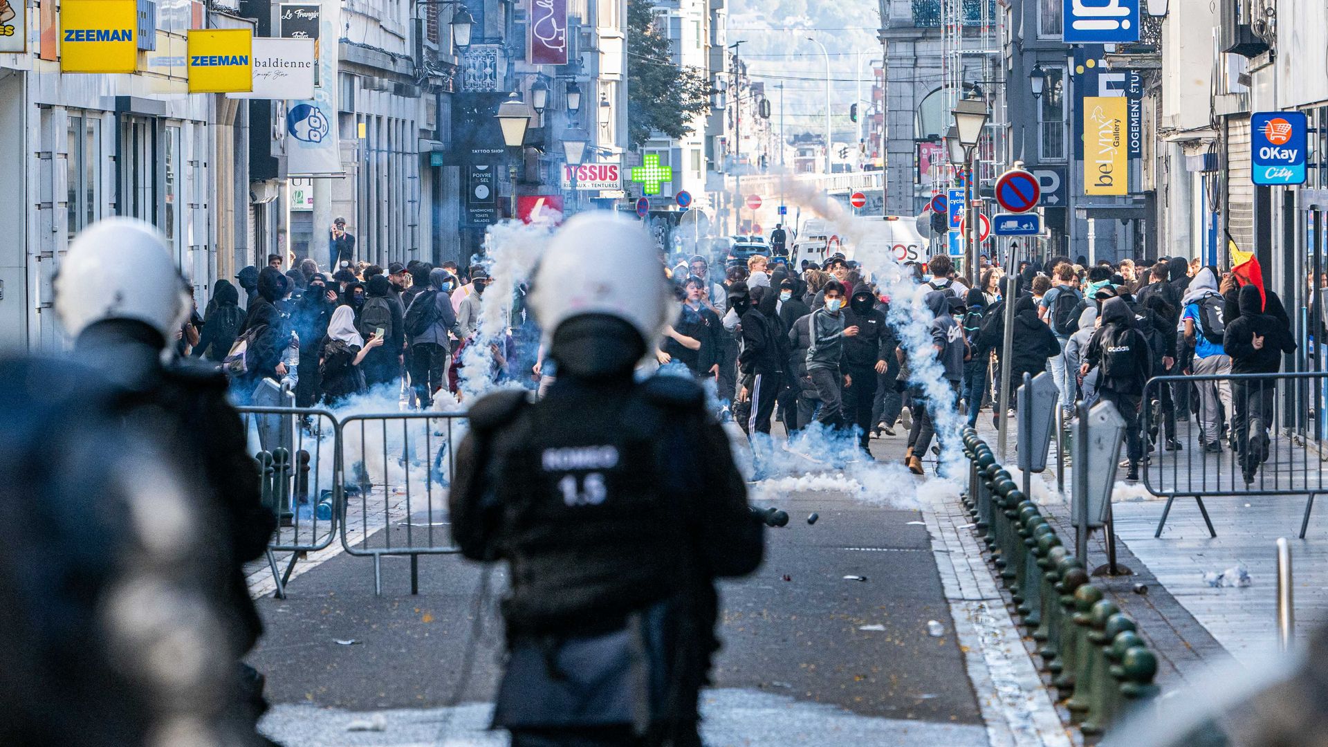 Crowded urban street protest seen from behind riot police in dark gear; smoke fills the road, barriers line the street, pedestrians populate the sidewalks and storefront signs loom on both sides.