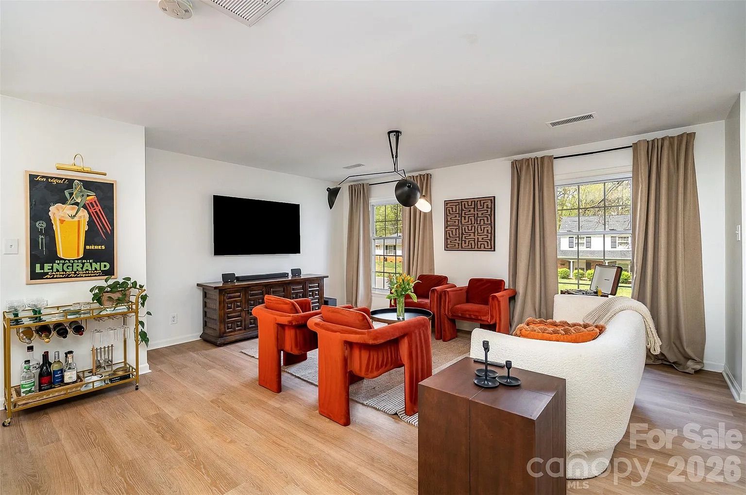 Modern living room with white walls, light wood floor, four orange chairs around a small round table, a white curved sofa, large windows with beige curtains, and a wall-mounted TV.