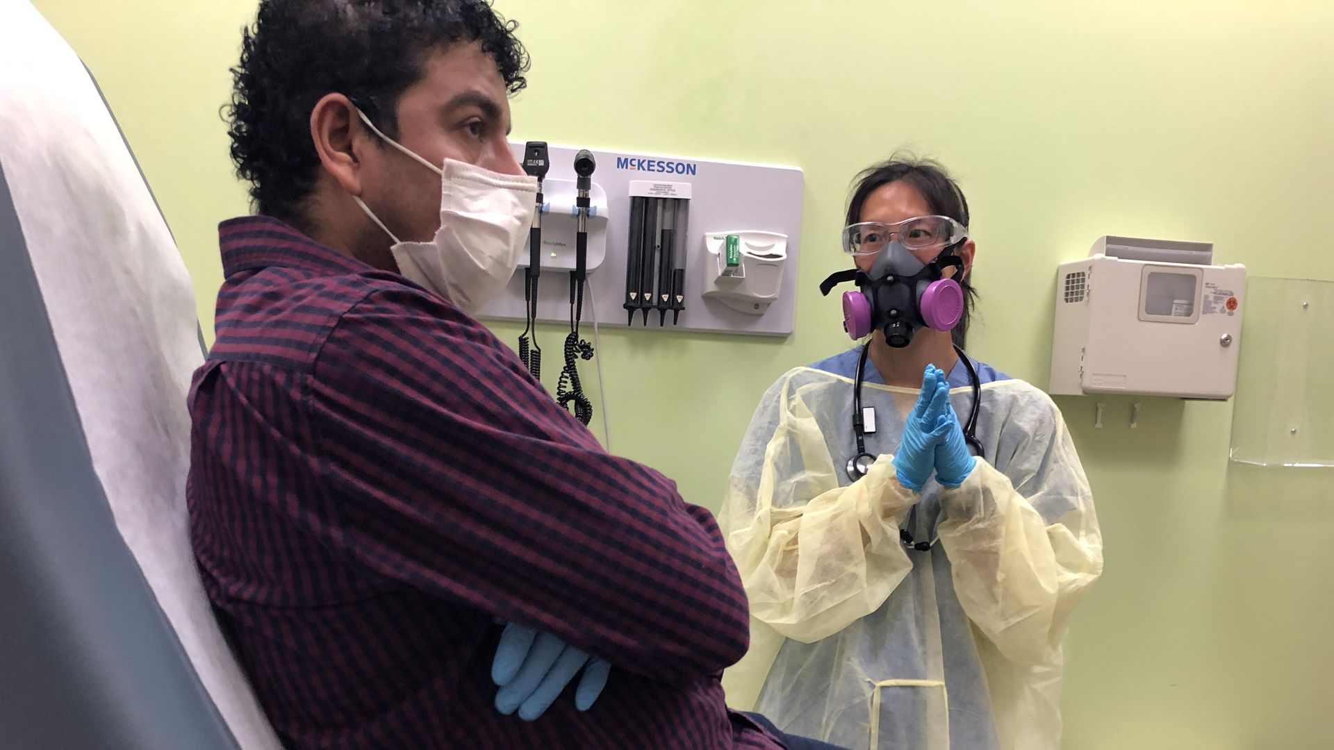 A physician assistant wears a mask and gown while talking to a patient who is sitting in an exam chair.