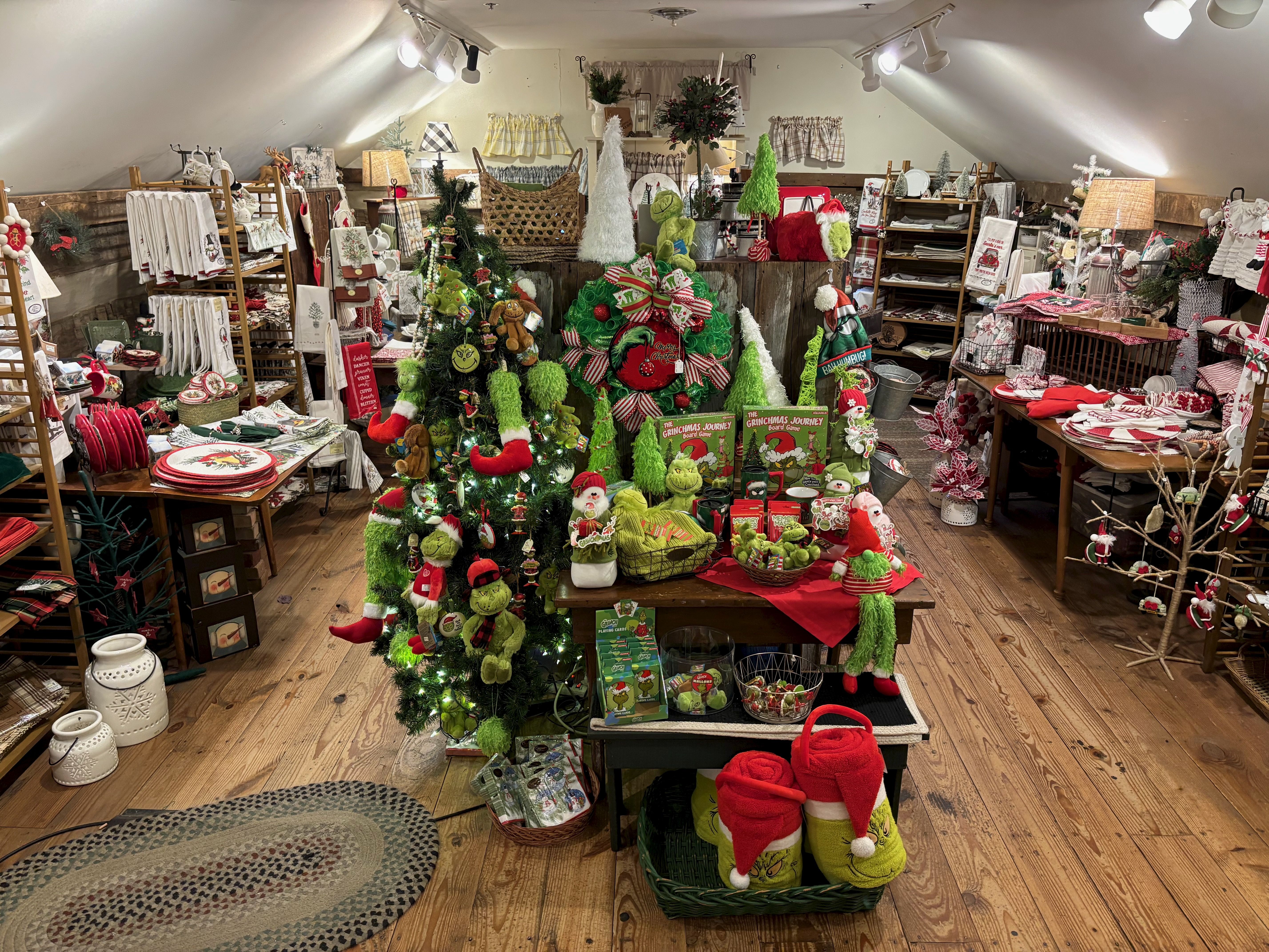 Cozy attic shop filled with Christmas decorations featuring Grinch-themed plush toys, wreaths, games, towels, and holiday textiles in red, green, and white colors under warm lighting.