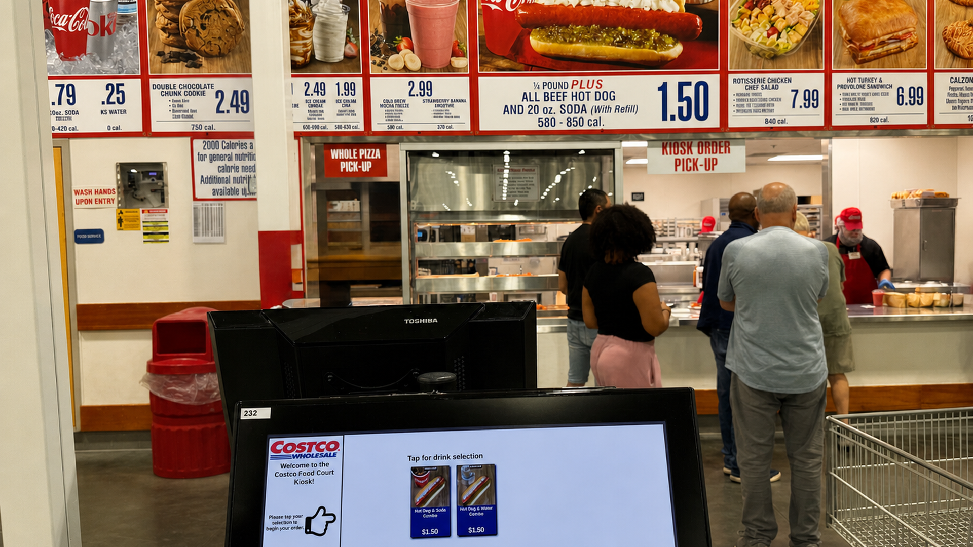 Inside a Costco food court, bold menu boards line the back wall with photos of pizzas, hot dogs, drinks, and sandwiches. Customers wait in line near a black Toshiba monitor at the checkout area.