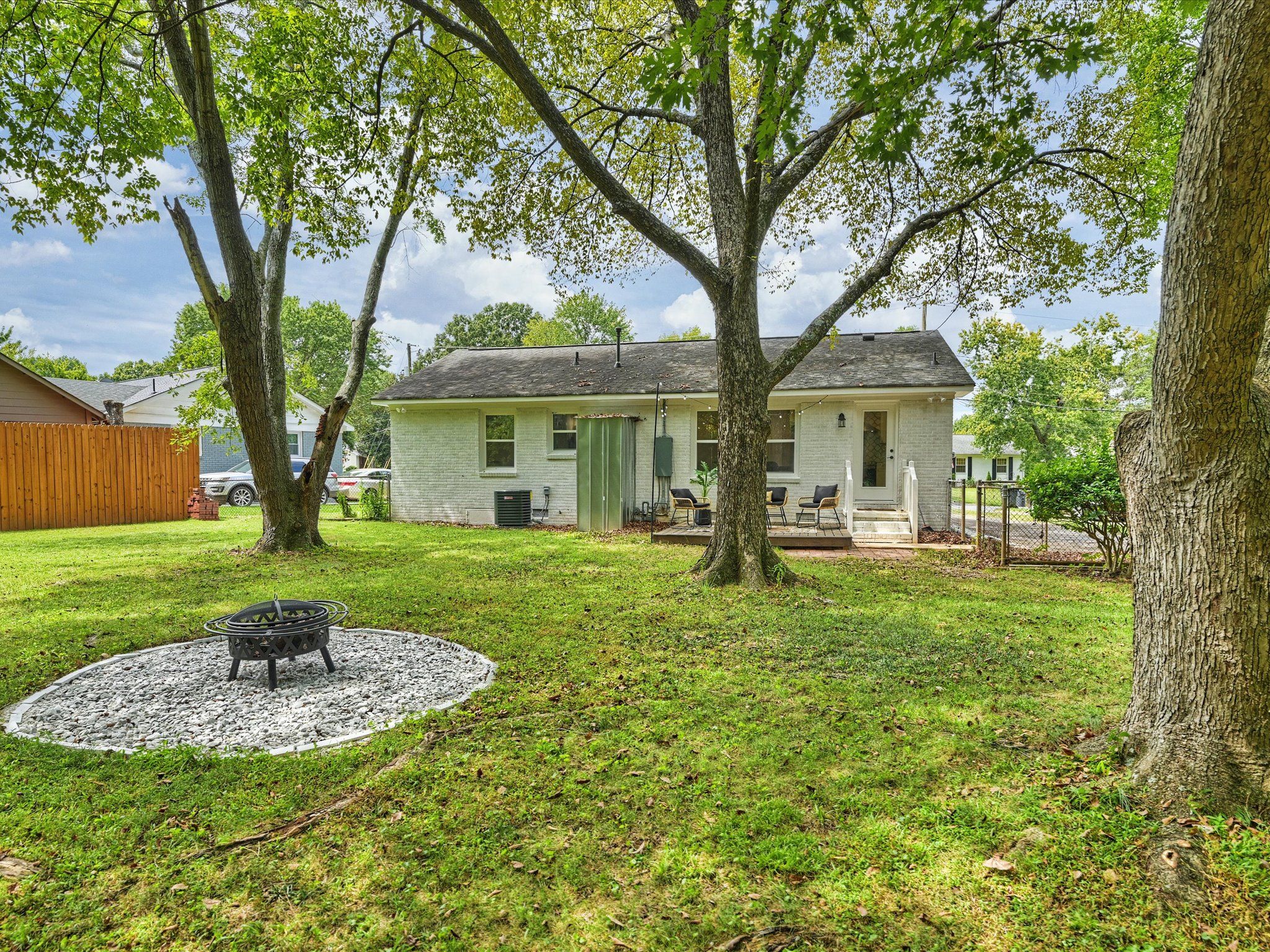 Backyard view of a white brick house with a small deck, patio chairs, three large trees, green grass, and a black metal fire pit on a circular gravel bed.