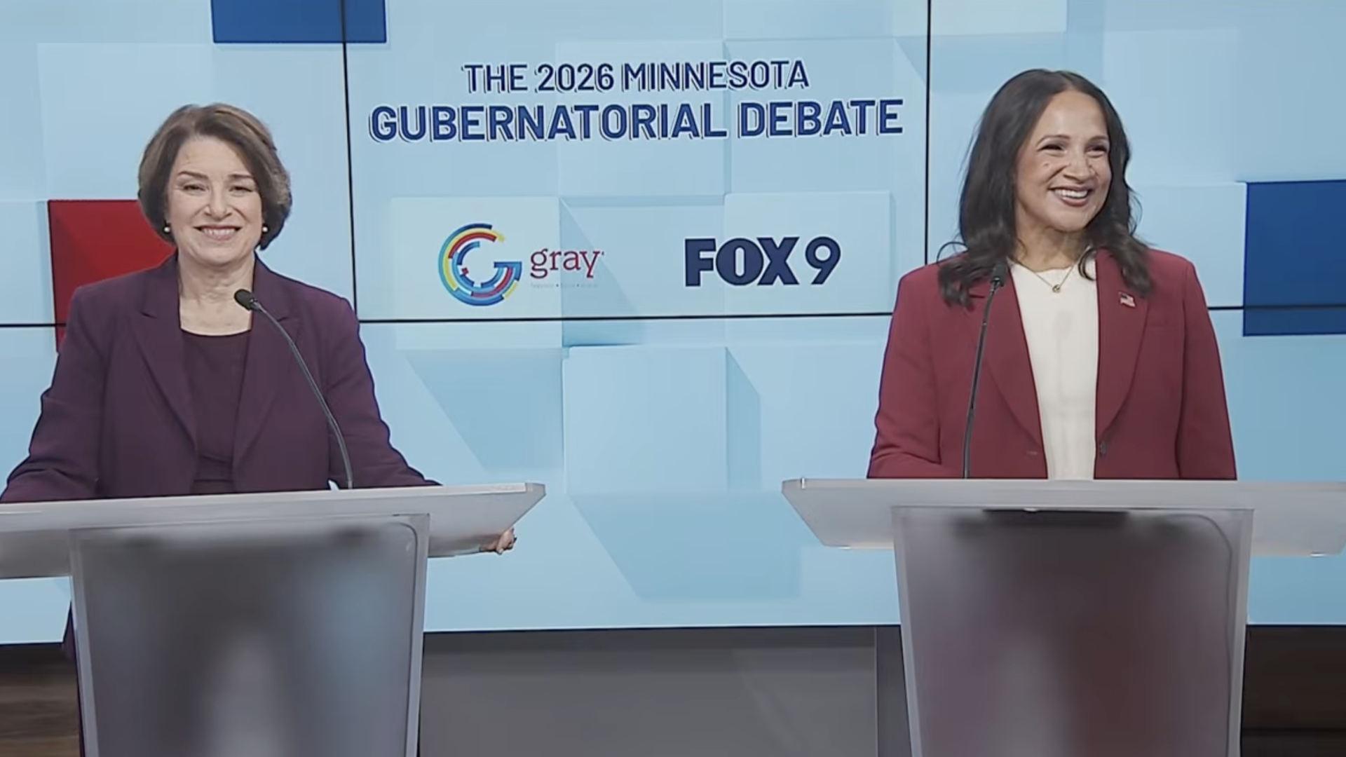 Two women stand at podiums during the 2026 Minnesota Gubernatorial Debate; left in a purple suit, right in a red blazer, backdrop displays the event title and logos.