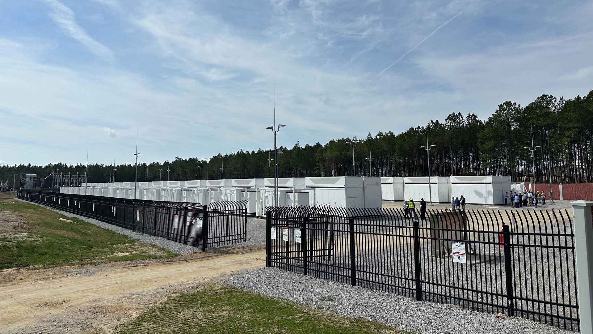 Secure industrial site with rows of white container-like units behind black metal fencing. A group of people and workers in helmets stand near the units. Trees line the background.