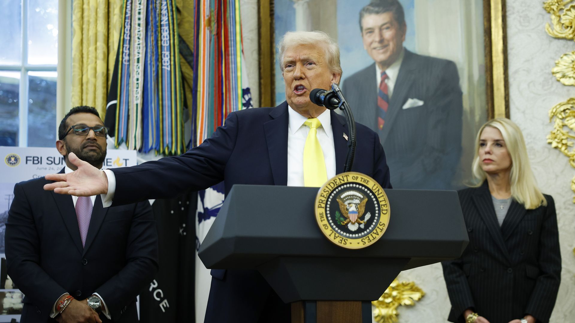 President Trump wears a yellow tie while speaking in the Oval Office. Pam Bondi stands to his right, and Kash Patel stands to his left. 