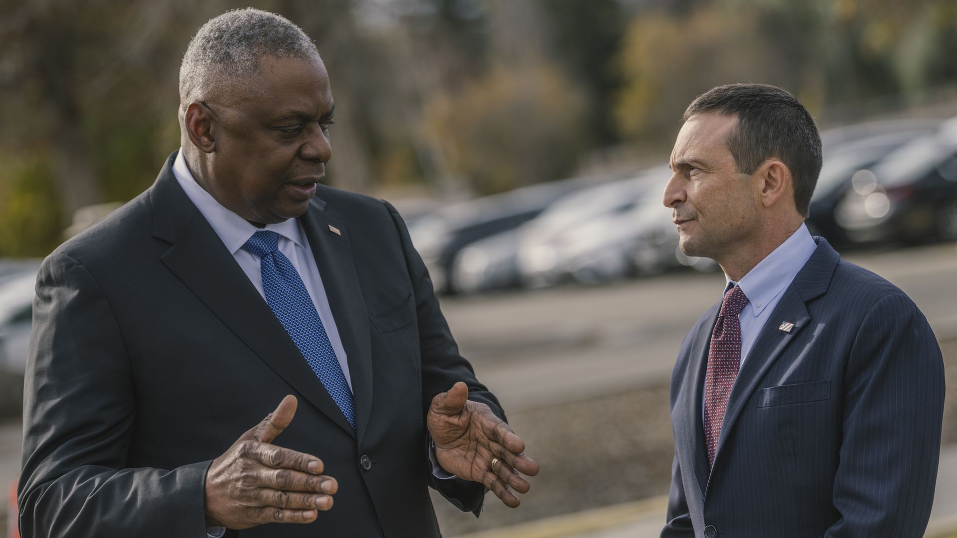 Two men, Lloyd Austin and Doug Beck, talk in a parking lot. Both are wearing suits, one with a blue tie, the other red. Austin is gesturing as he speaks, while Beck listens.