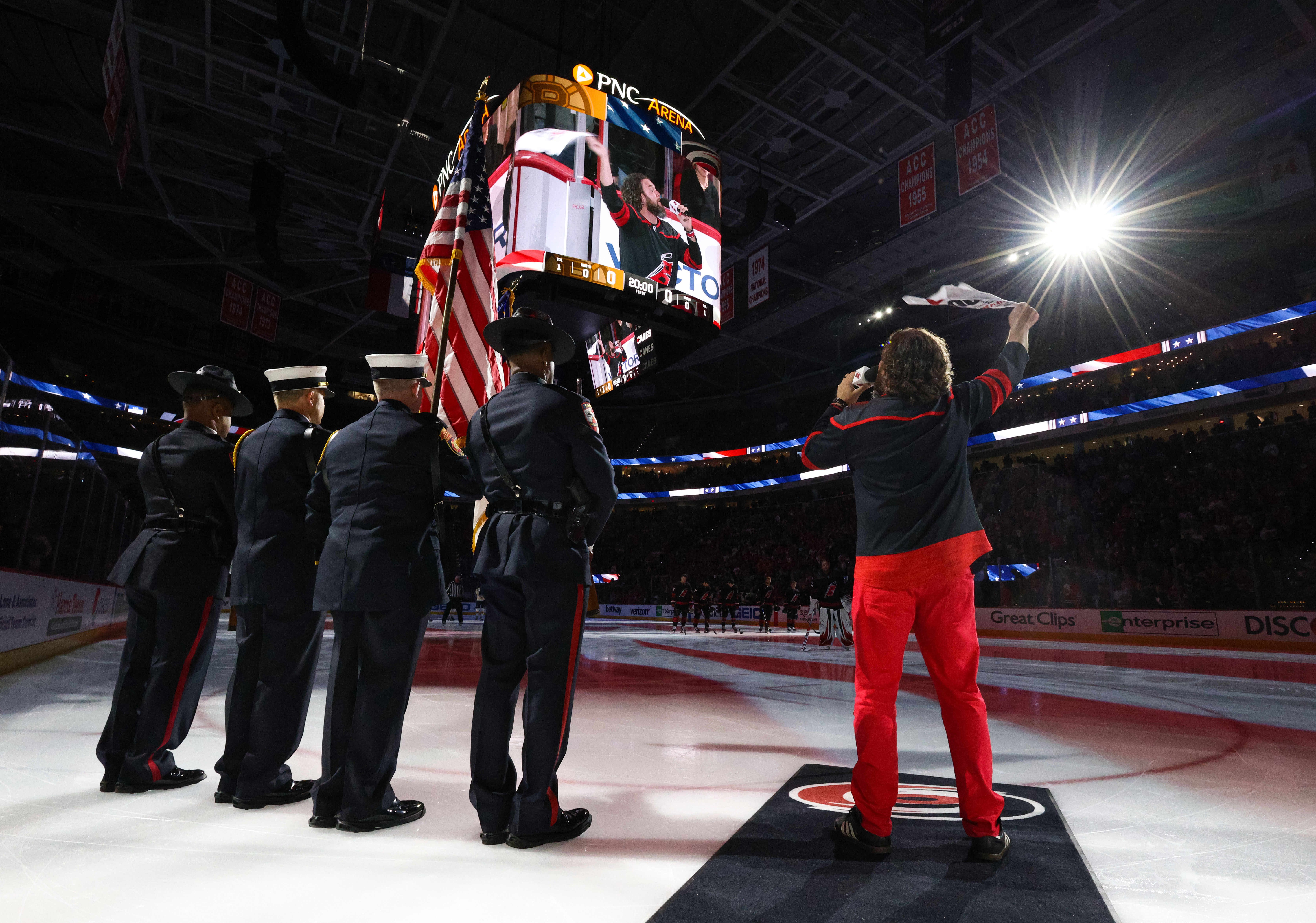 Adam Lee Decker sings the National Anthem on the ice prior to a 2022 Carolina Hurricanes playoff game in Raleigh. We see him from behind, wearing red pants and a black jersey and waving a towel. His face is on the jumbotron. Four soldiers beside him stand holding the American flag.
