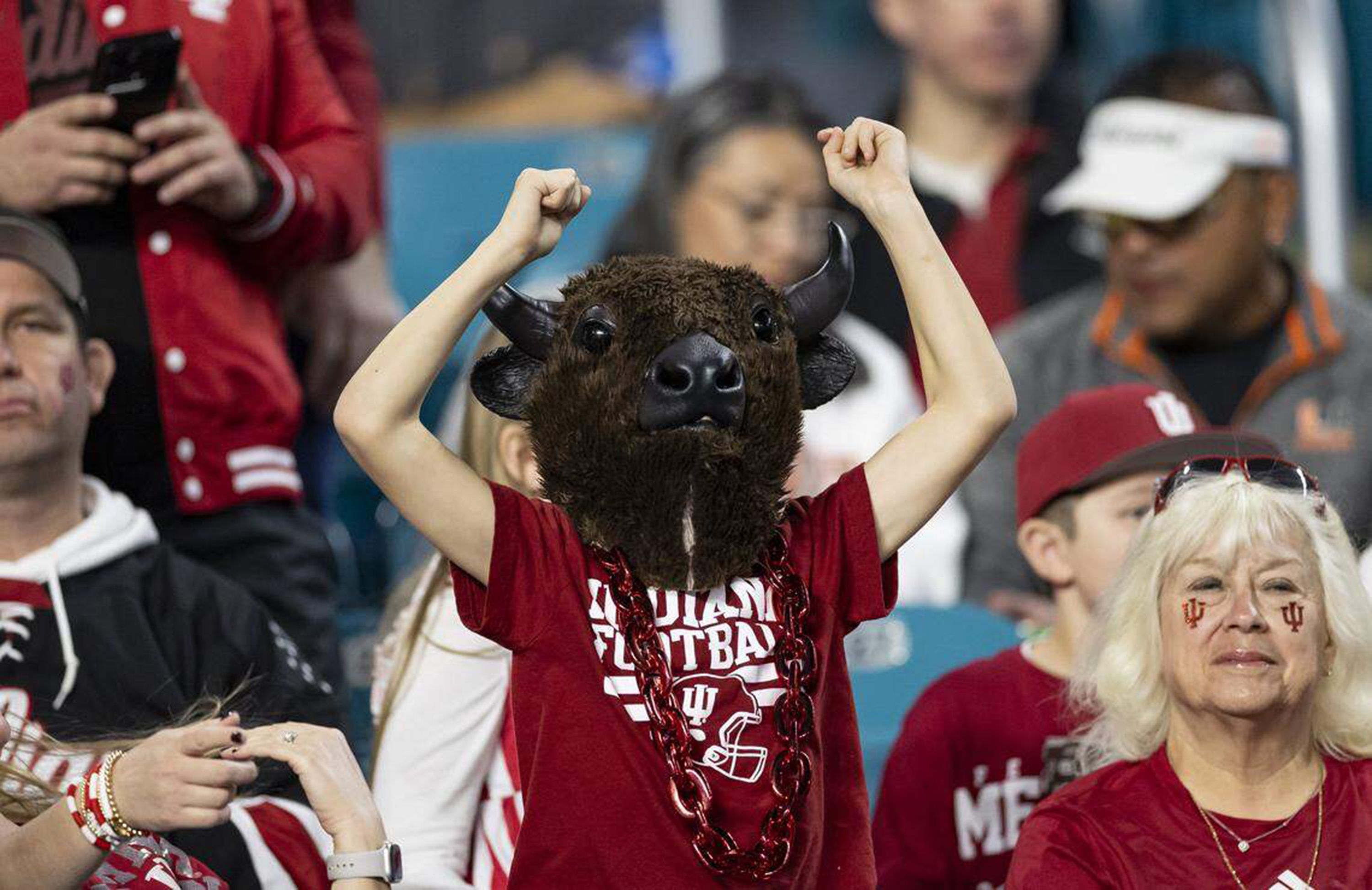 Fan wearing a large bison head mask, red Indiana Football shirt, and red chain necklace cheering with raised fists in a crowd at a sports event.