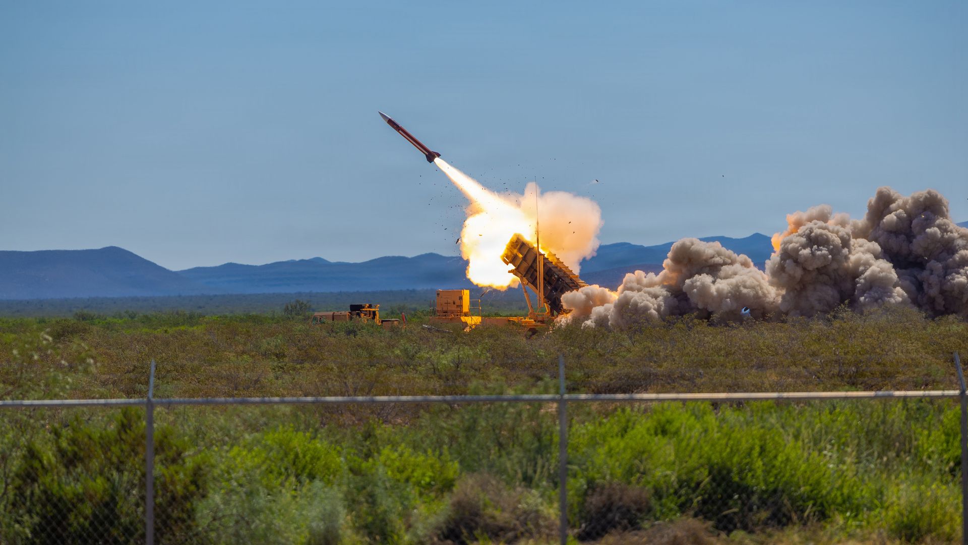 Blue sky over a desert launch site as a red rocket blasts upward from a tilted launcher, leaving a bright flame and smoke plume.
