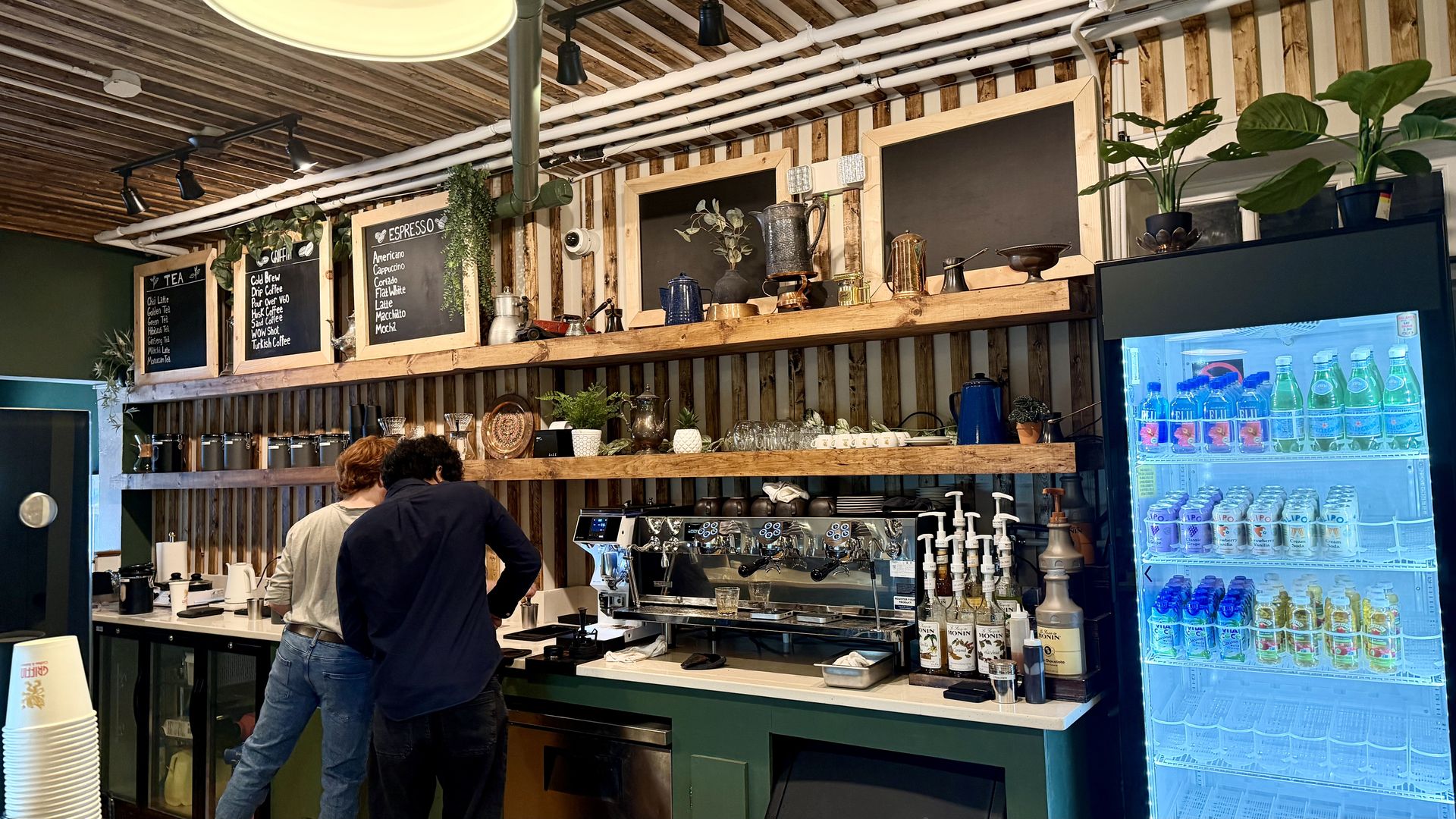 Two people working behind the counter of a cozy coffee shop with wooden decor, blackboards listing tea and espresso options, a coffee machine, syrups, plants, and a lit fridge with drinks.