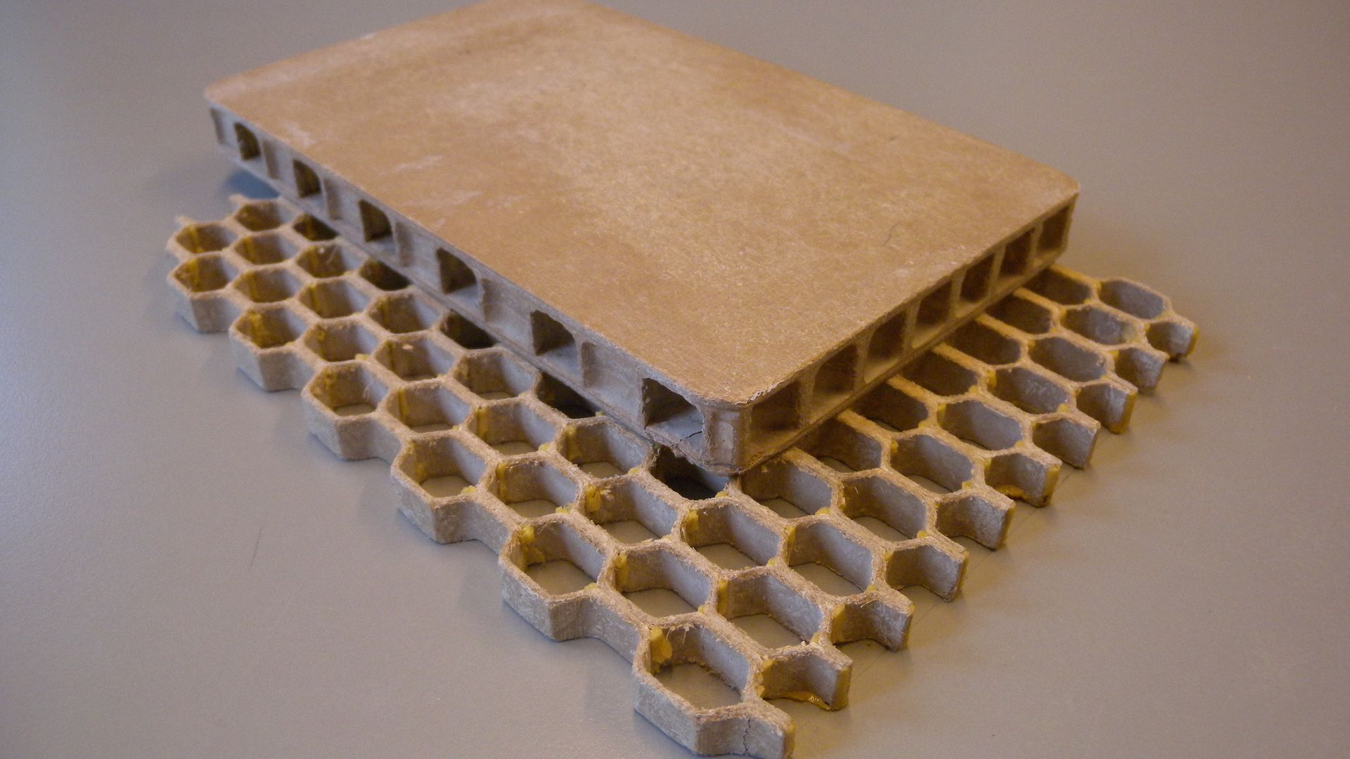 A photo shows two rectangular panels made from Moxy's Cerlos building material, which it produces from forest and farm scrap. One is a honeycomb panel, the other on top of it is flat and rectangular. Both are Earth-toned in hue.