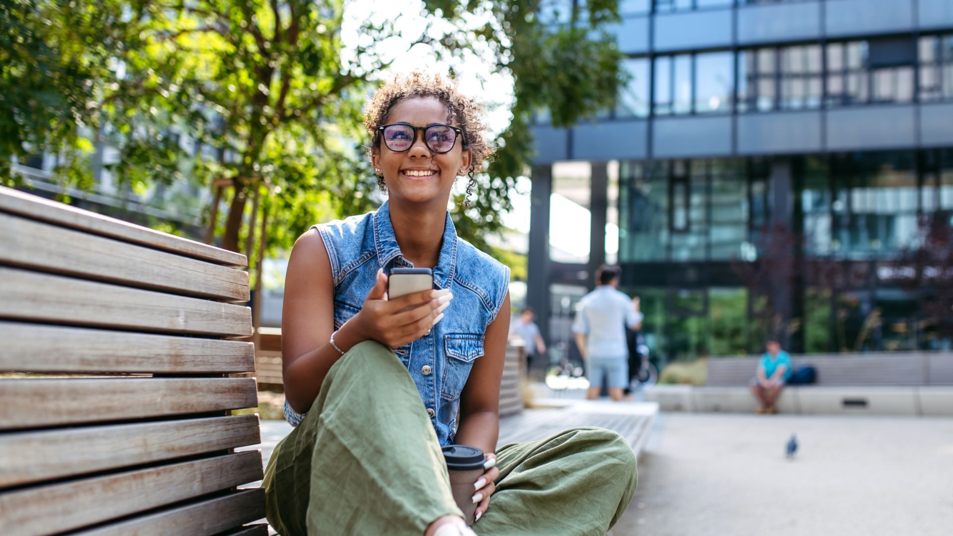 Young woman with curly hair and glasses sits on a wooden bench outside a modern glass building, smiling at her phone, wearing a denim vest and olive pants, coffee cup on her lap.