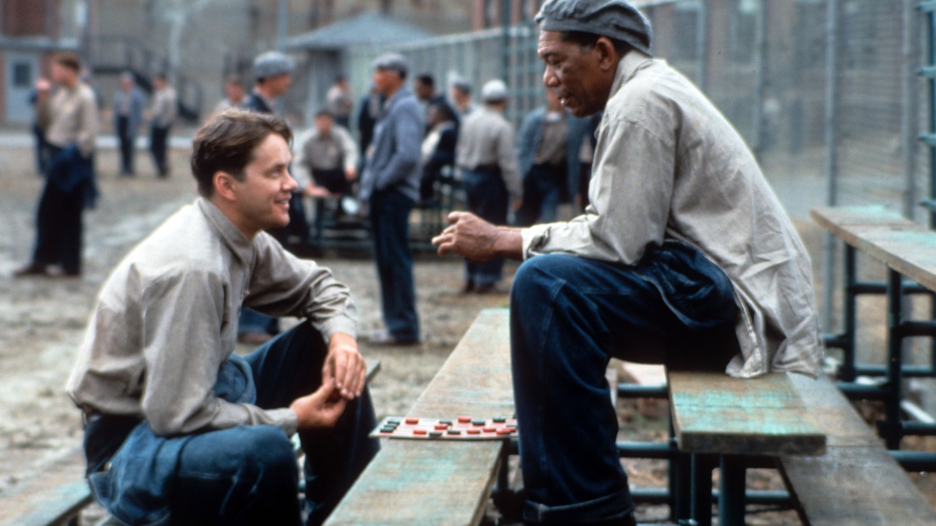 Two men in prison uniforms play checkers on weathered wooden benches in a prison yard, surrounded by other inmates in gray and beige clothing, with chain-link fencing in the background.