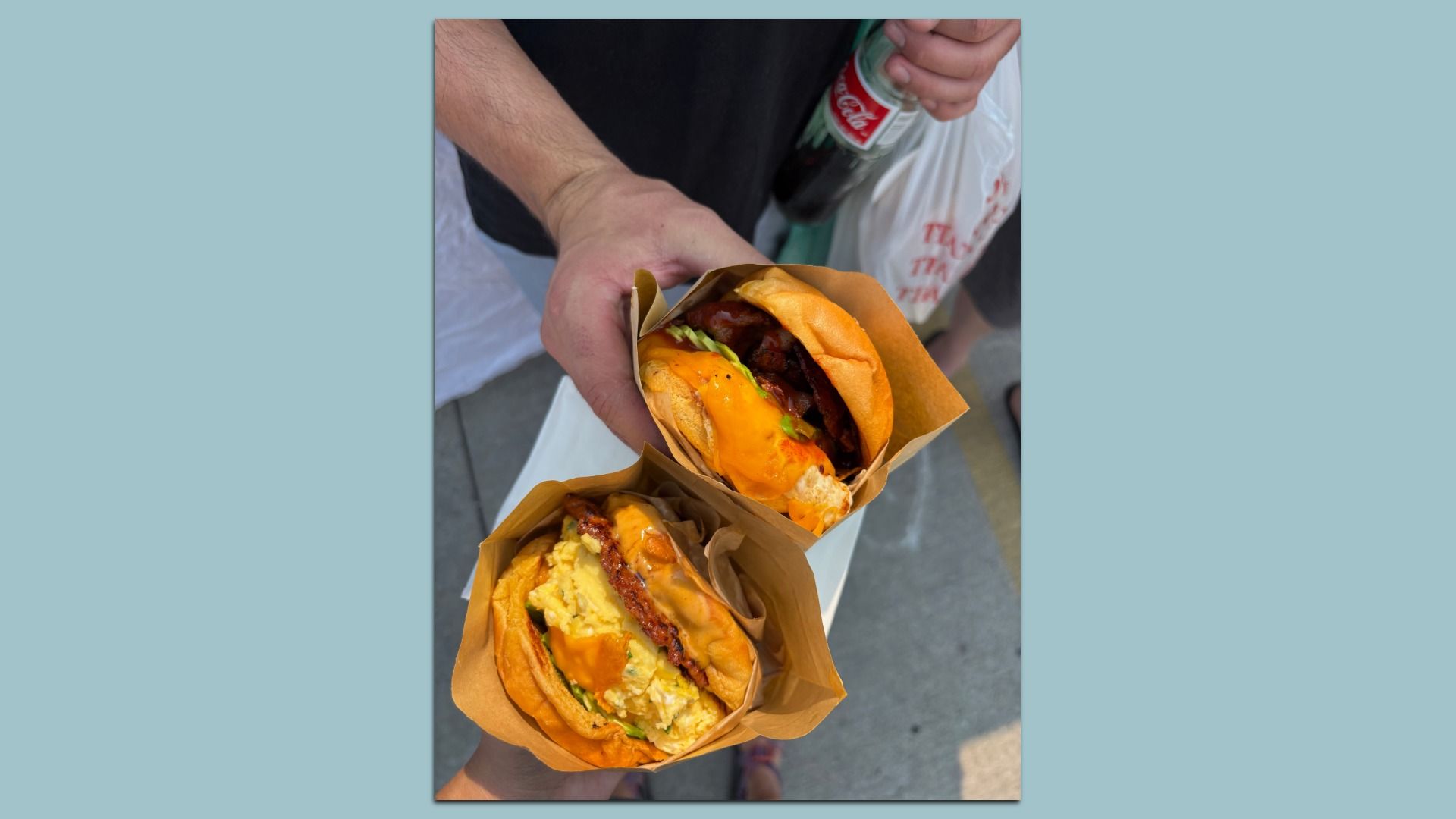 Two breakfast sandwiches in brown wrappers with melted cheddar and lettuce, held by a person in a black shirt; one hand grips a Coca-Cola bottle and a white plastic bag.