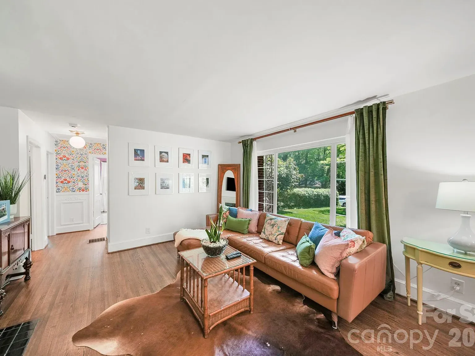 Bright living room with white walls, a tan leather sofa with colorful cushions, a glass coffee table on a brown cowhide rug, a large window with green curtains, and a gallery wall of framed photos.