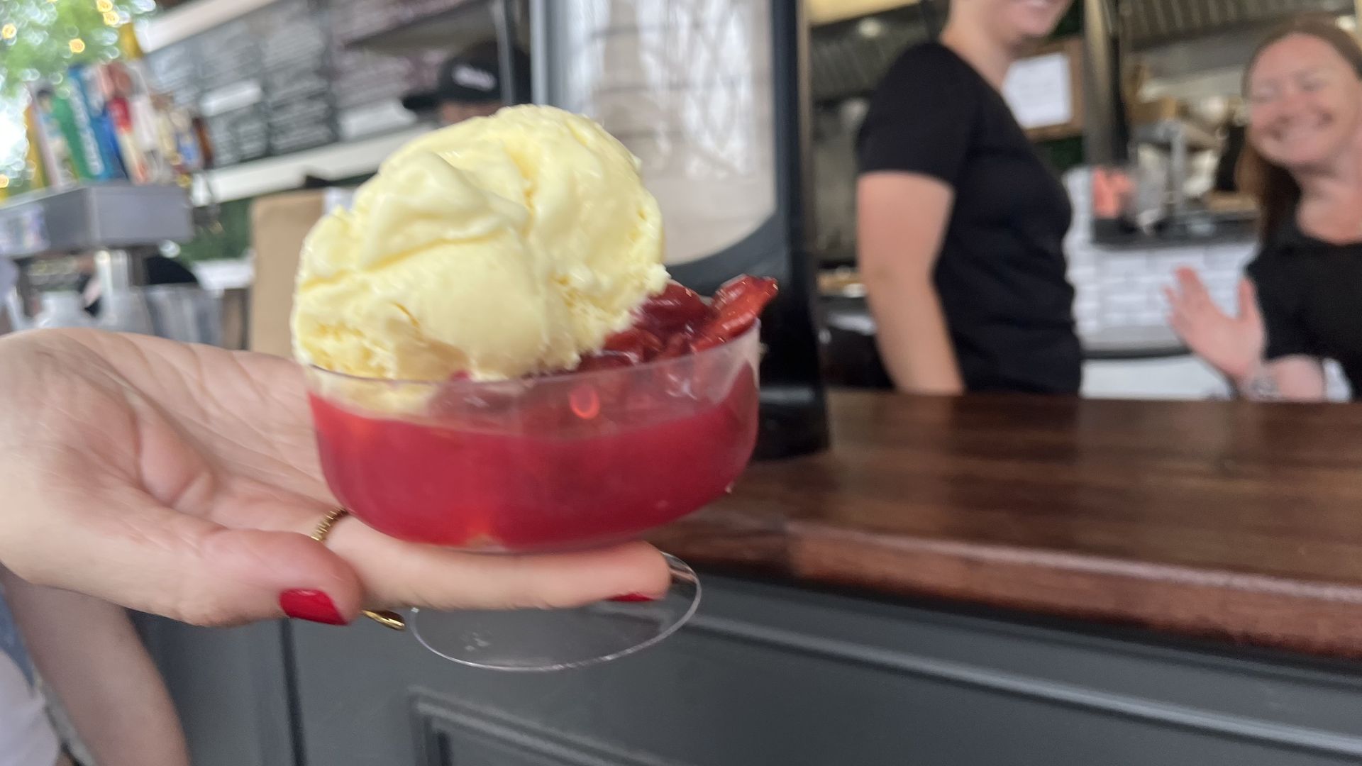 A hand with red nail polish holds a glass of bright red drink topped with a large pale yellow scoop of ice cream; blurred cafe counter and staff visible in the background.