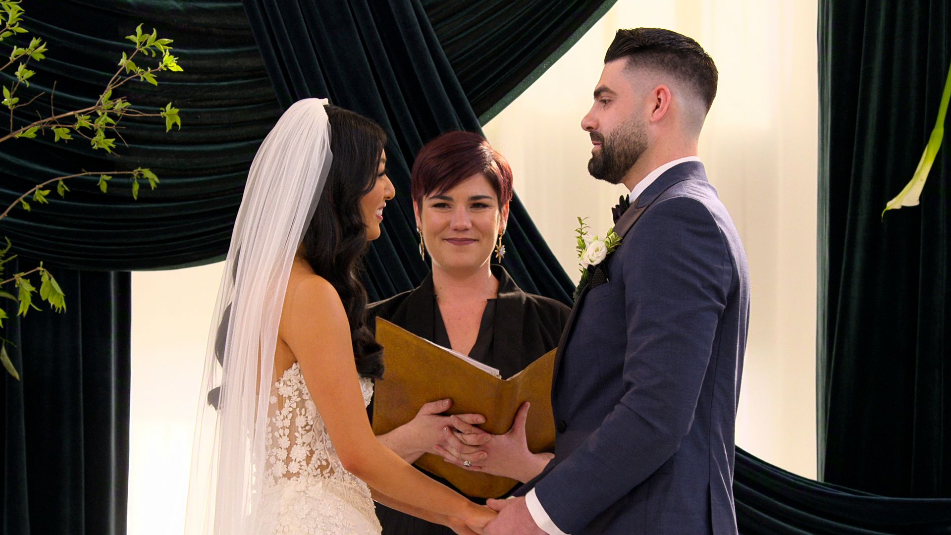 Bride in white lace dress and veil smiling at groom in dark blue suit holding hands during wedding ceremony with officiant holding book in background with dark drapes and greenery.