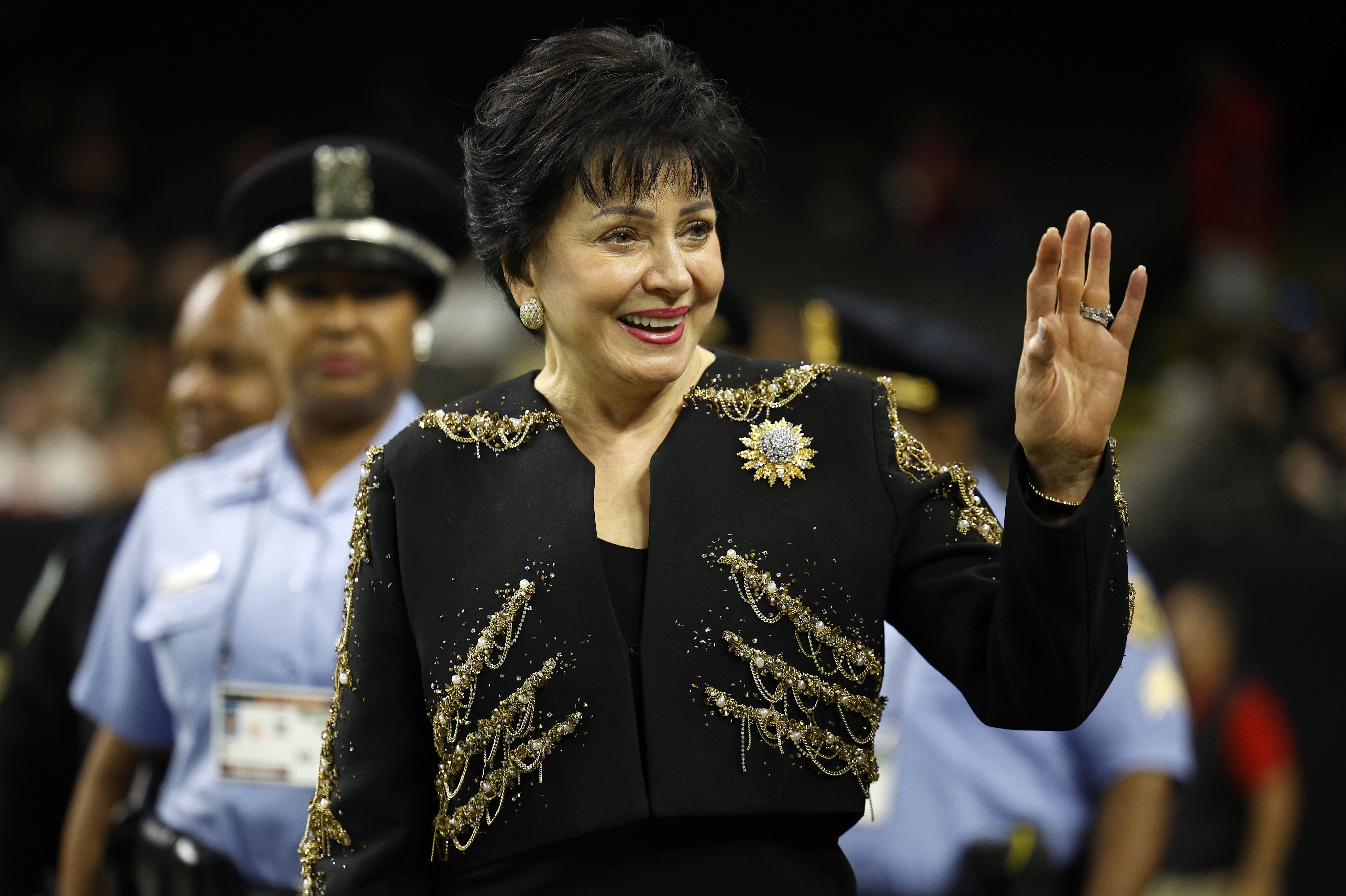 Gayle Benson wears a black and gold jacket and waves.