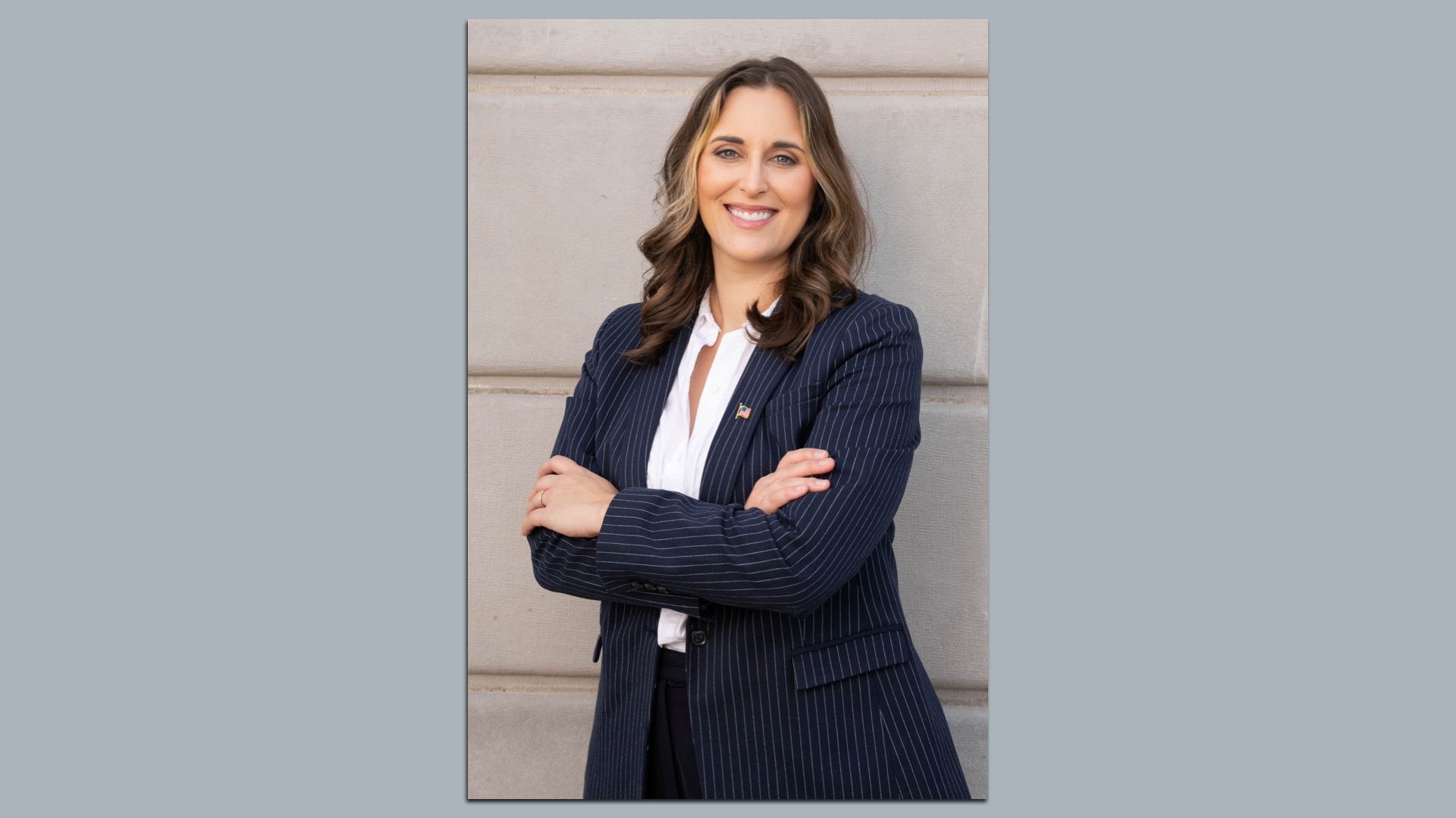 Woman with wavy brown hair, wearing a navy pinstripe blazer and white blouse, arms crossed and smiling, standing against a light stone wall.