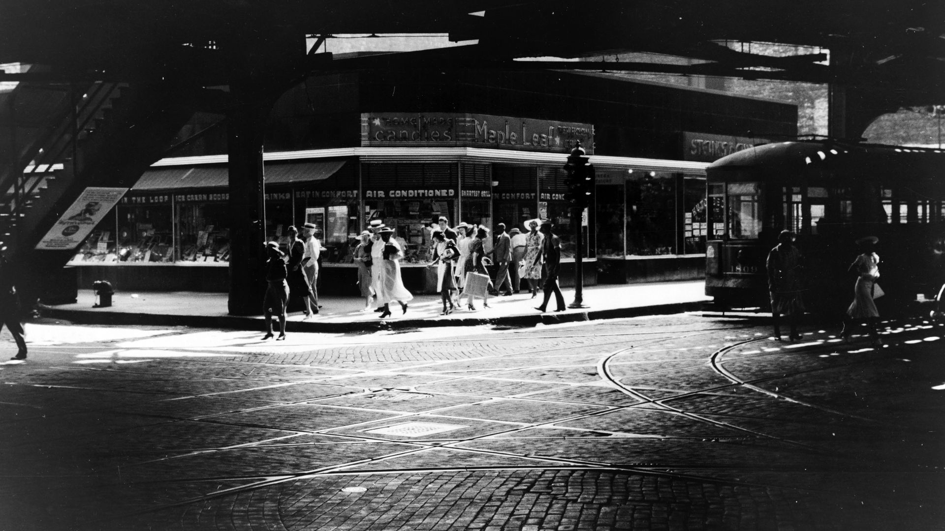 Black-and-white street scene beneath an elevated structure; a crowd walks past storefronts with signs like "candles" and "Maple Leaf"; curved tram tracks cross the cobblestones.
