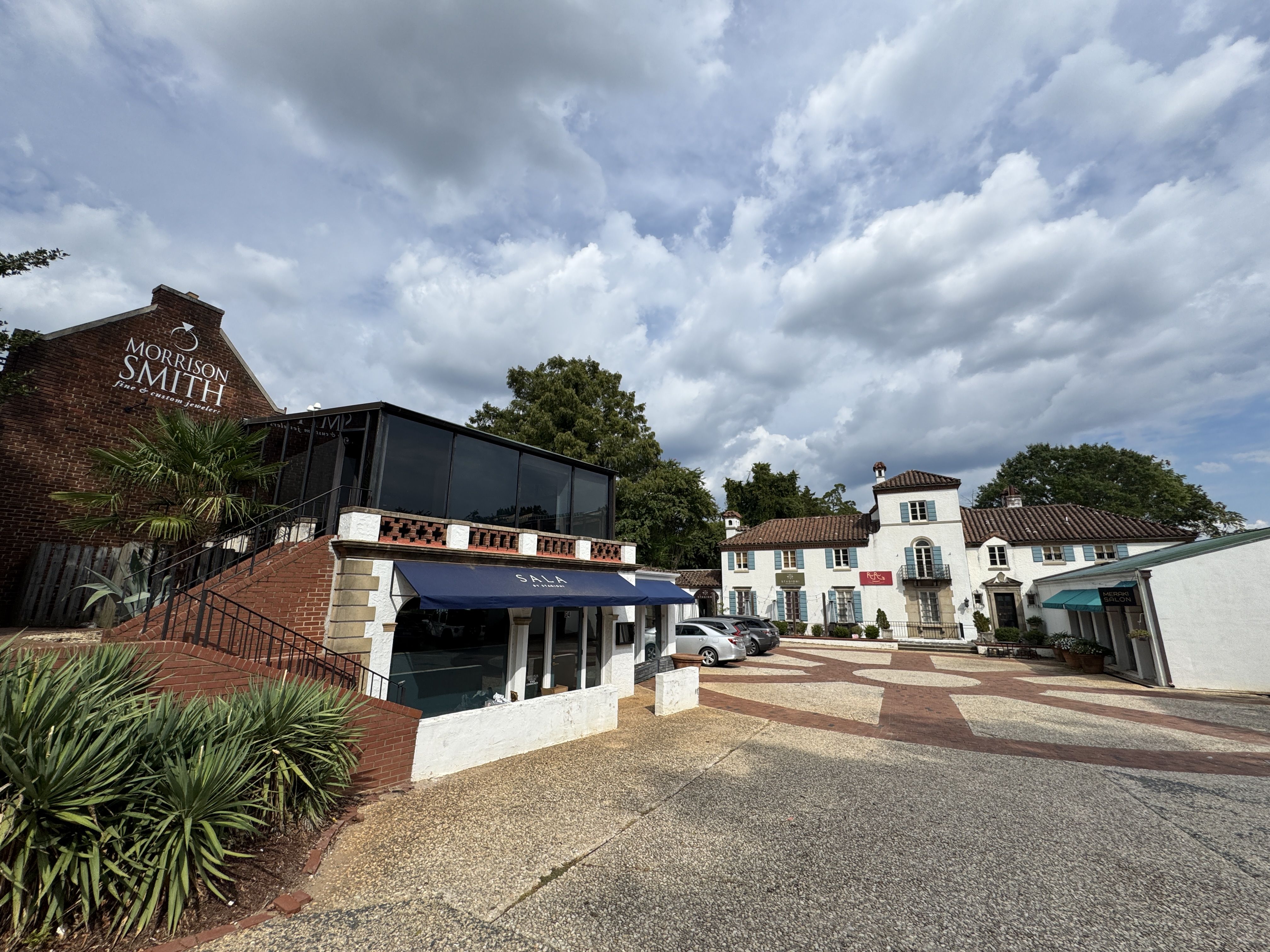Outdoor view of a plaza with a brick building labeled Morrison Smith and a white building labeled Red Rooster and Salon under a partly cloudy sky.