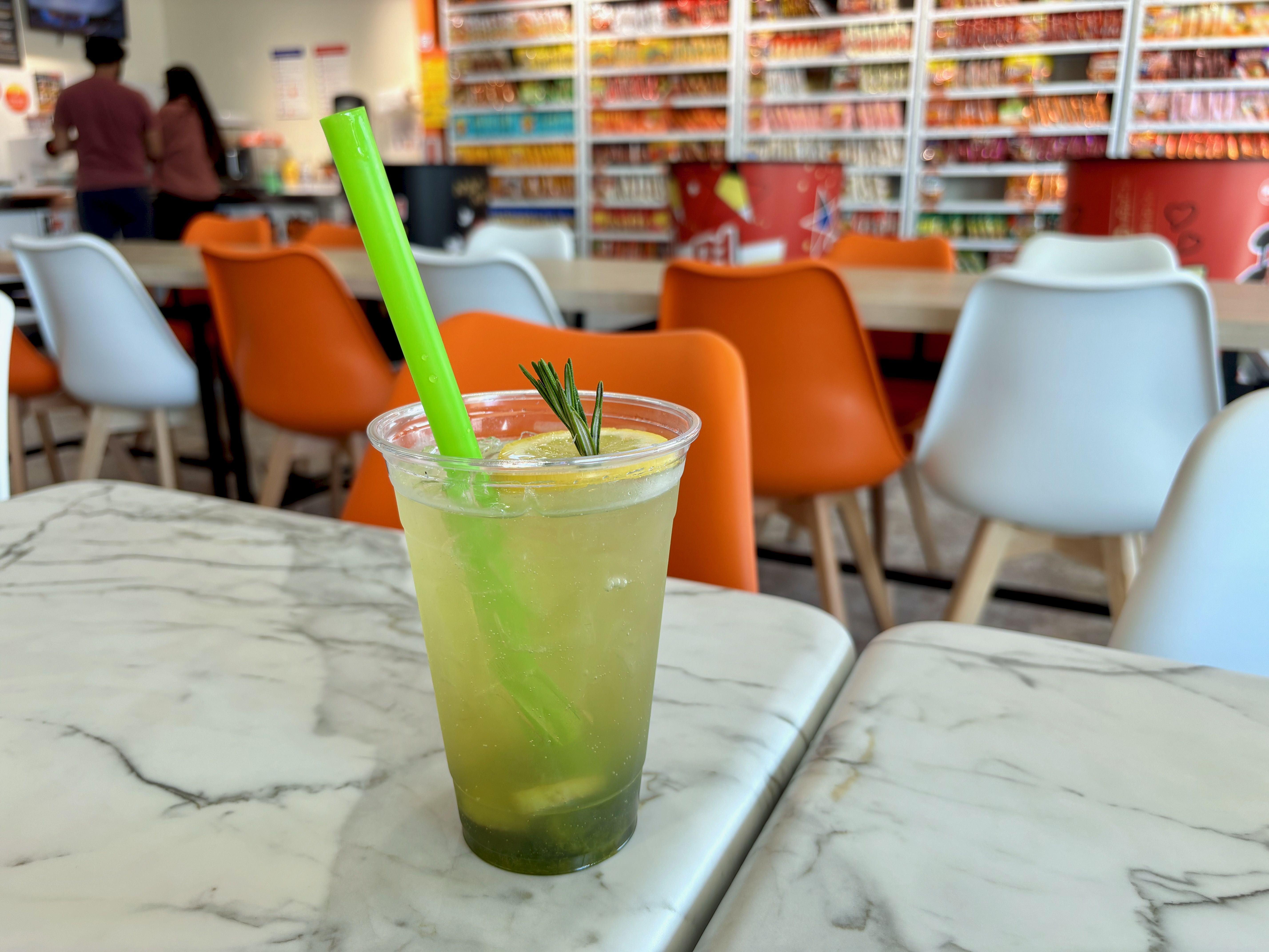 Close-up of a lime-green iced drink with a bright green straw and rosemary sprig on a marble table; blurred orange and white chairs and snack shelves in the background.