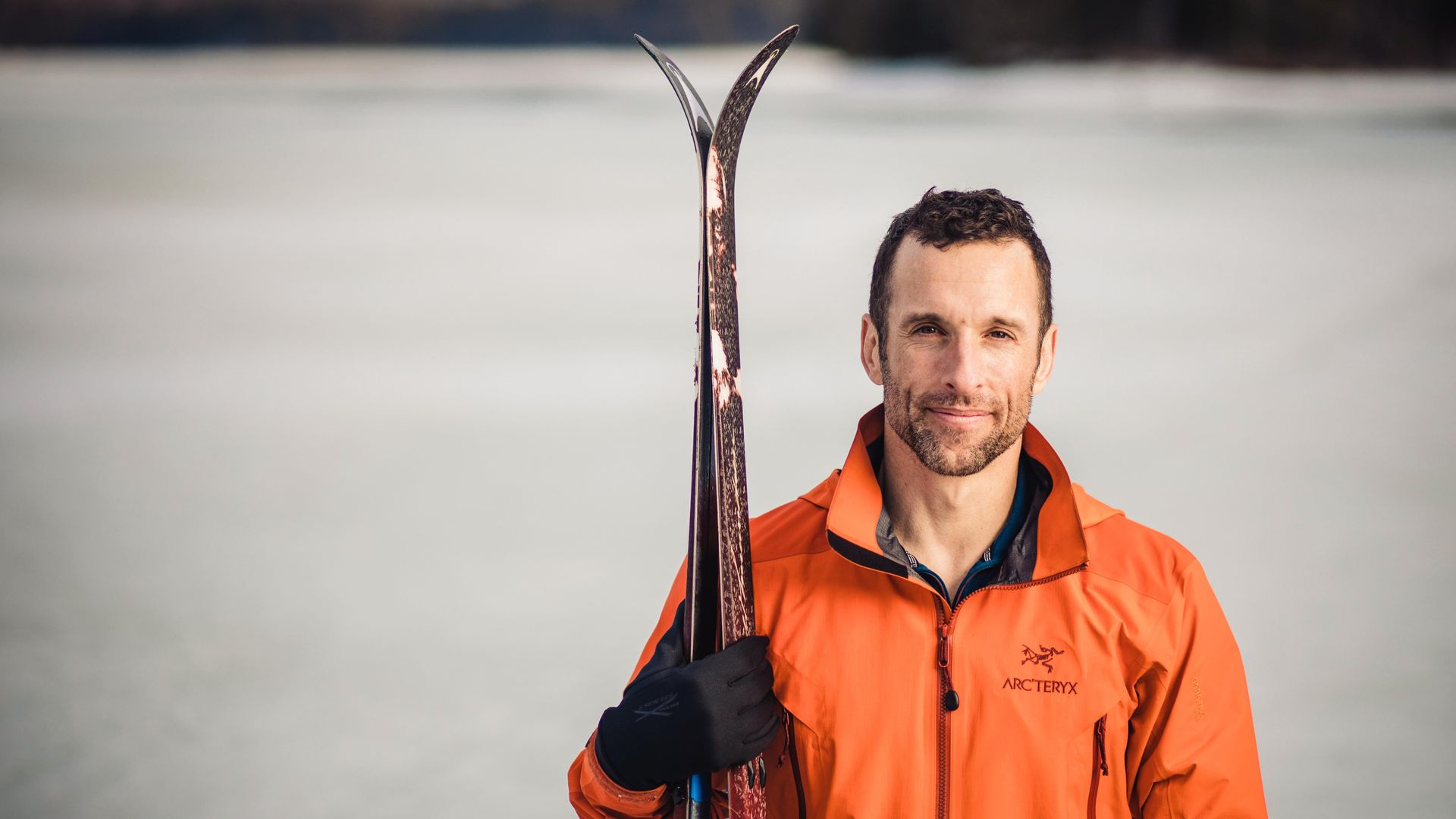 Sebastian Audy stands in an ice field with an orange jacket holding skis