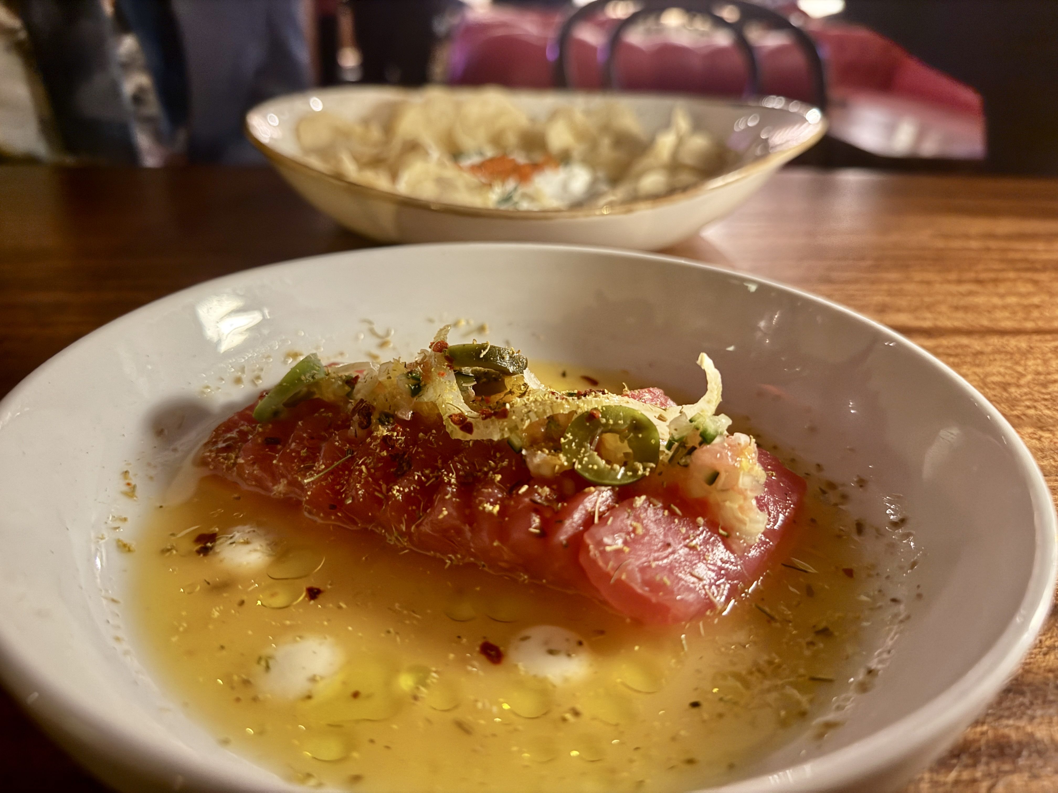 White bowl with a slice of raw tuna topped with sliced green chilies and spices in a light broth, on a wooden table with a blurred bowl of chips in the background.