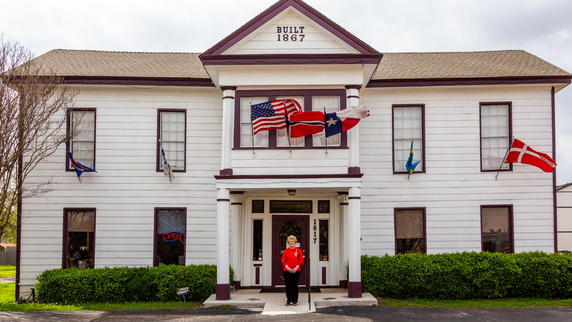 White two-story historic building with maroon trim and a peaked front gable that reads BUILT 1867. Flags hang from the balcony, including US and Texas flags. A woman in a red shirt stands at the entrance.