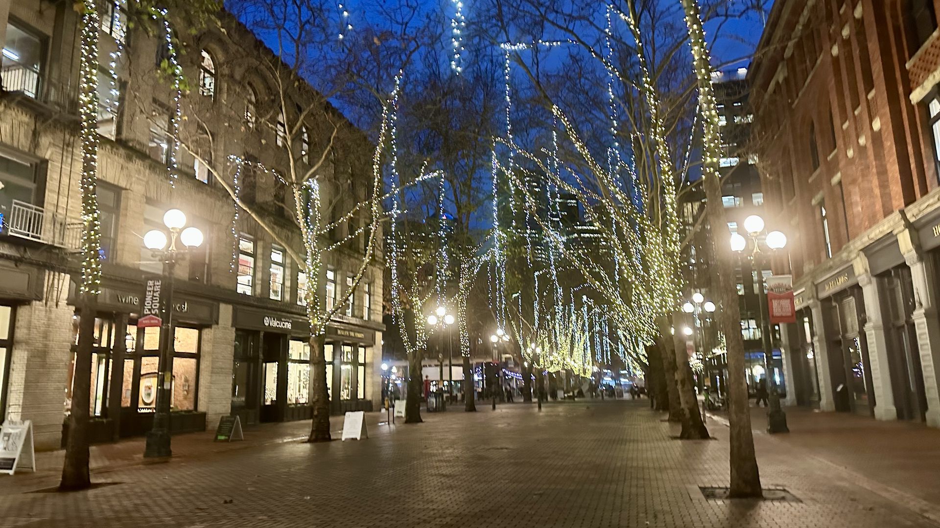 Lights drape from trees in Pioneer Square at night, with classic street lamps glowing and the brick pavement of the plaza reflecting the glow.